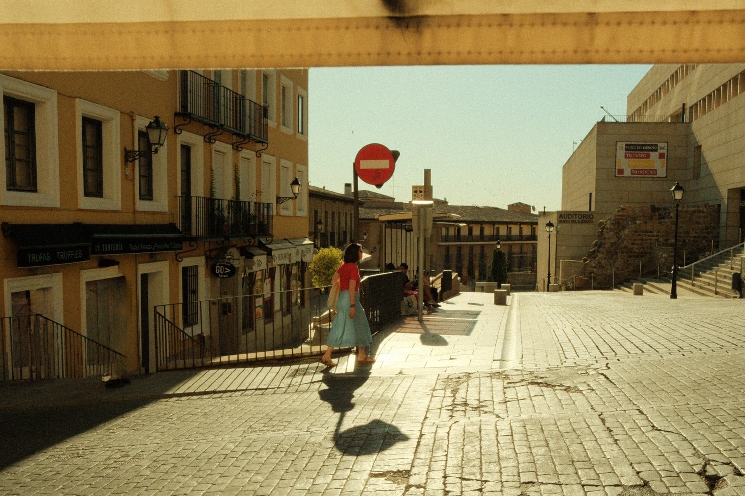 Woman in red top walking on a sunlit street in Toledo with a ‘No Entry’ sign and storefronts in the background