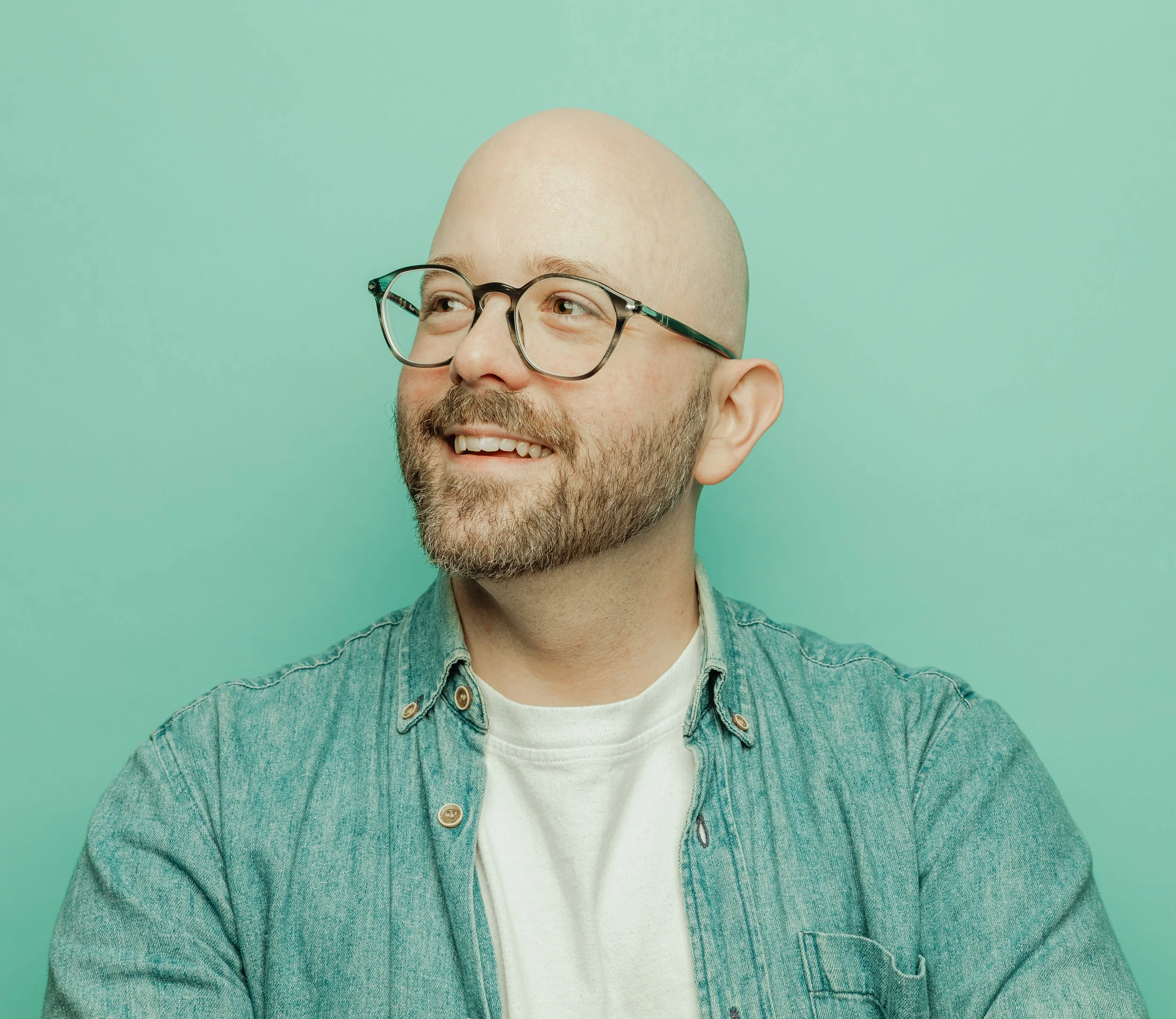 Smiling man with glasses and a beard wearing a denim shirt against a mint green background.