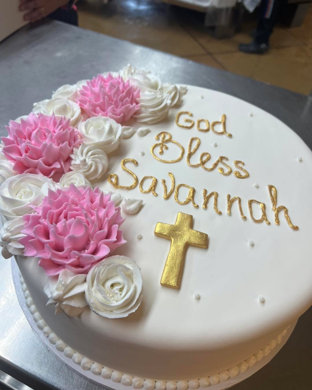 This is a beautifully decorated round cake with white fondant, adorned with pink and white buttercream flowers. The cake features the message “God Bless Savannah” written in gold icing, along with a gold cross and small pearl-like decorat