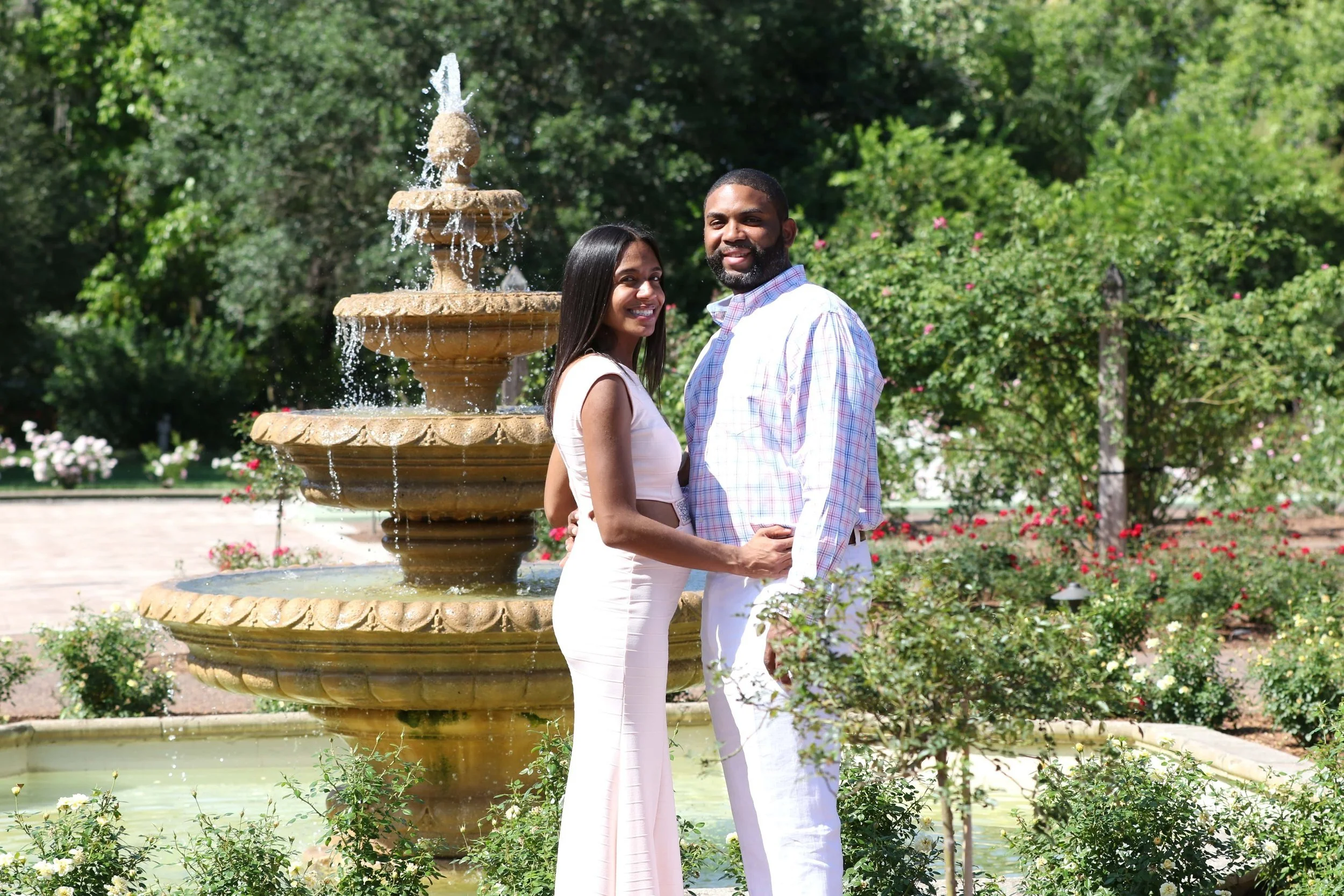 A smiling couple stands holding hands in front of a multi-tiered stone fountain in a garden, surrounded by greenery and blooming flowers.