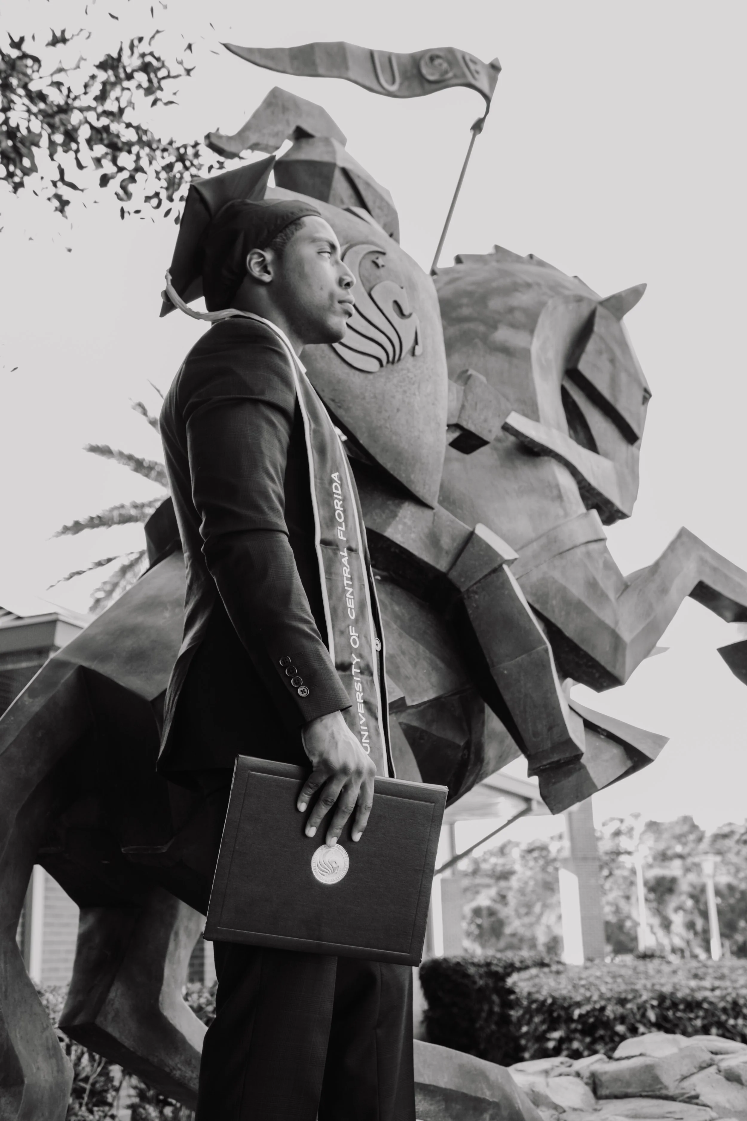 A young man in graduation cap and gown with a university sash stands outdoors holding a diploma, with a large bronze statue of a lion and a horse behind him.