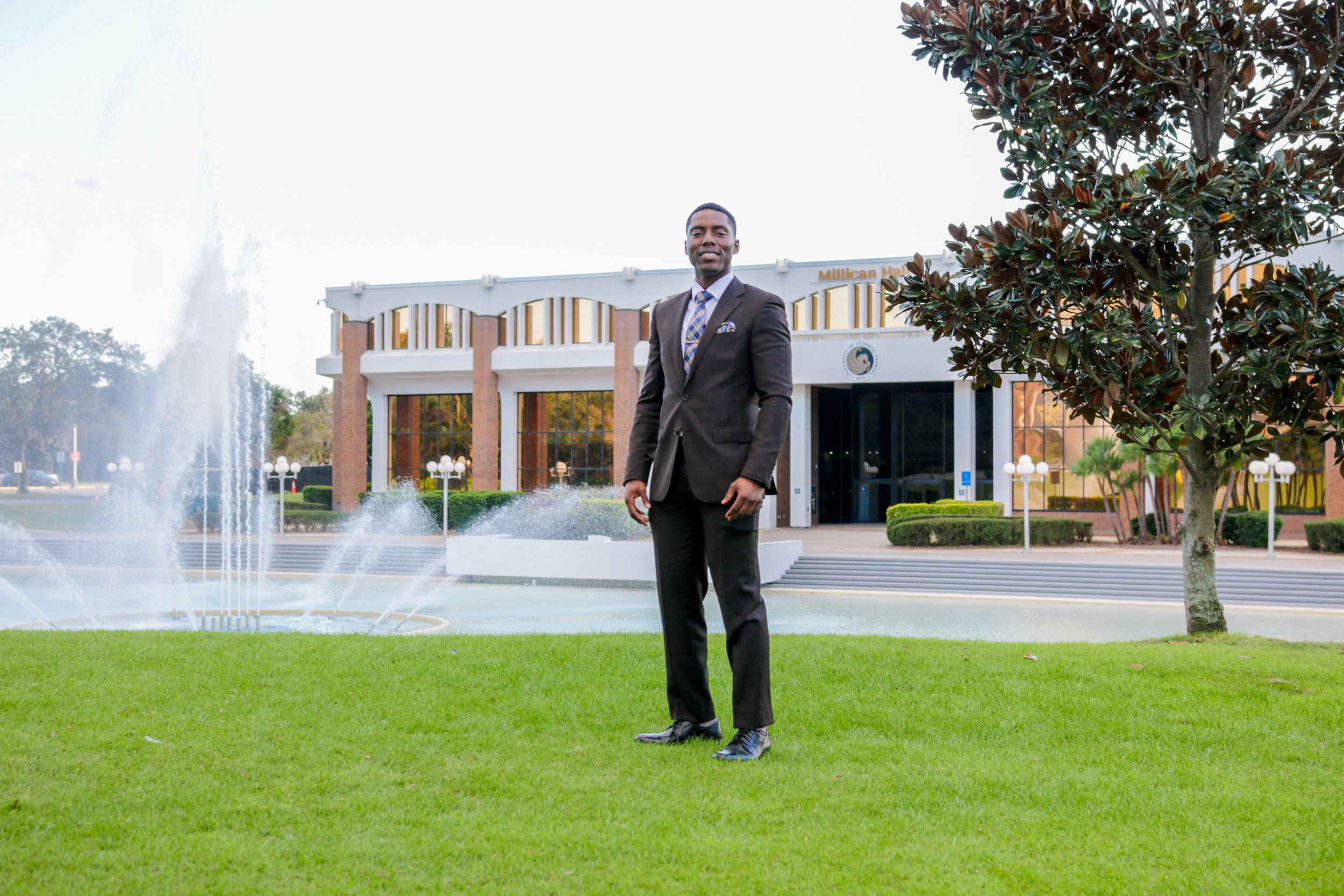 A man in a black suit and tie standing on a well-maintained grassy area in front of a modern building with large glass windows, a fountain, and a tree to the right.
