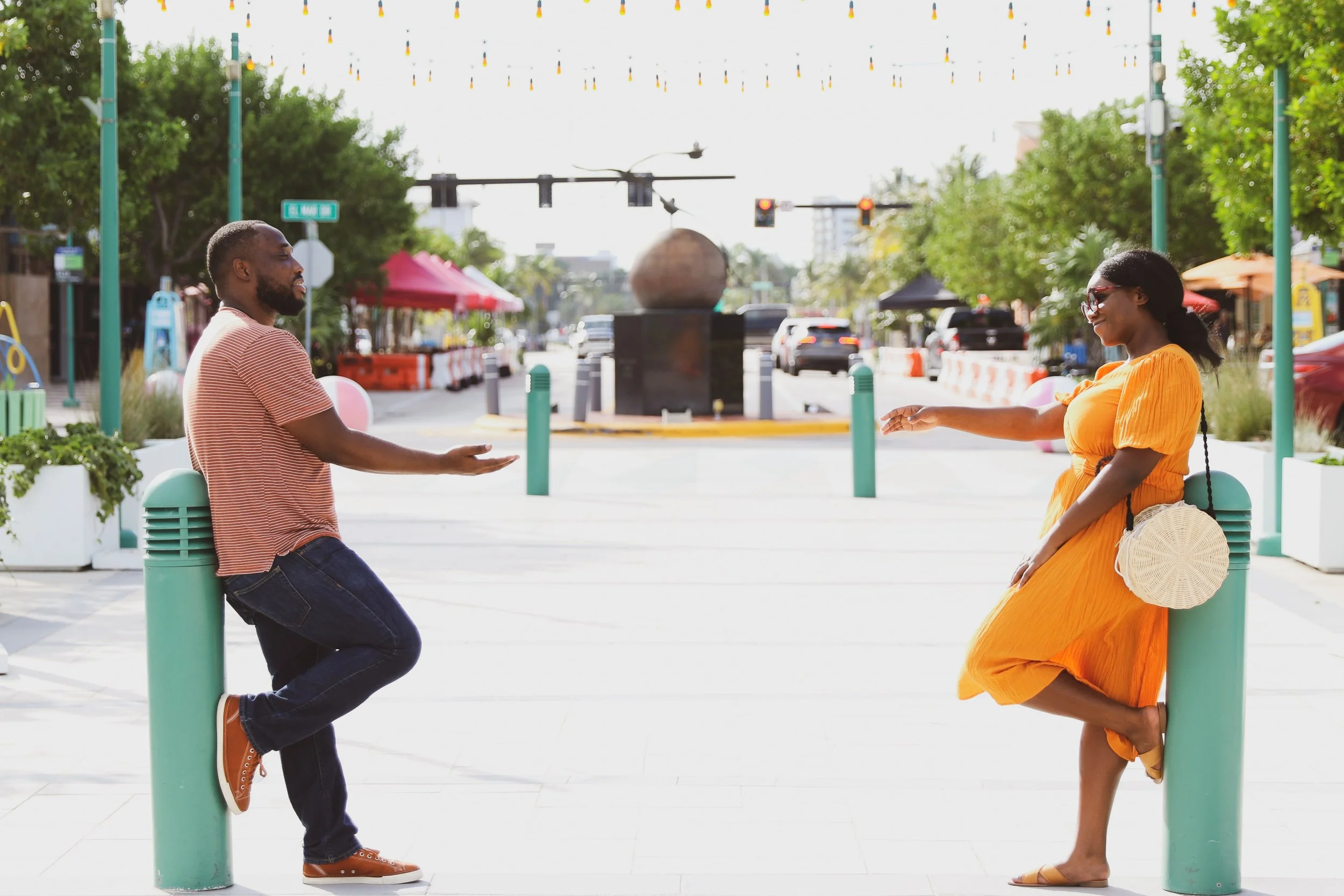 A man and woman are standing on opposite sides of a street, reaching towards each other in a friendly greeting or gesture. The man leans on a green post while the woman stands with one foot crossed over the other, leaning on a similar post. They are 