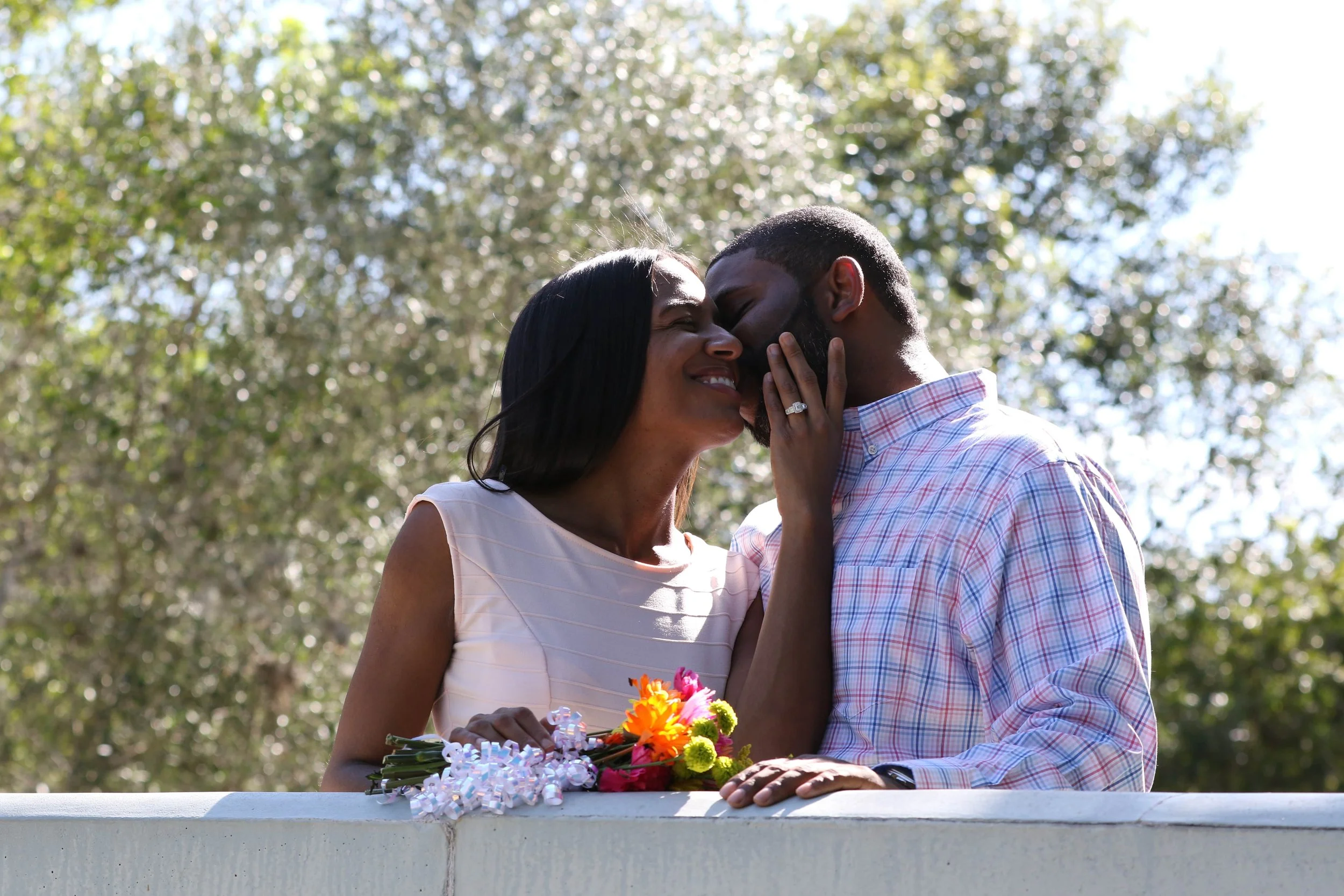 A romantic couple sharing a kiss outdoors, with a woman holding a bouquet of flowers and smiling while a man gently touches her face.