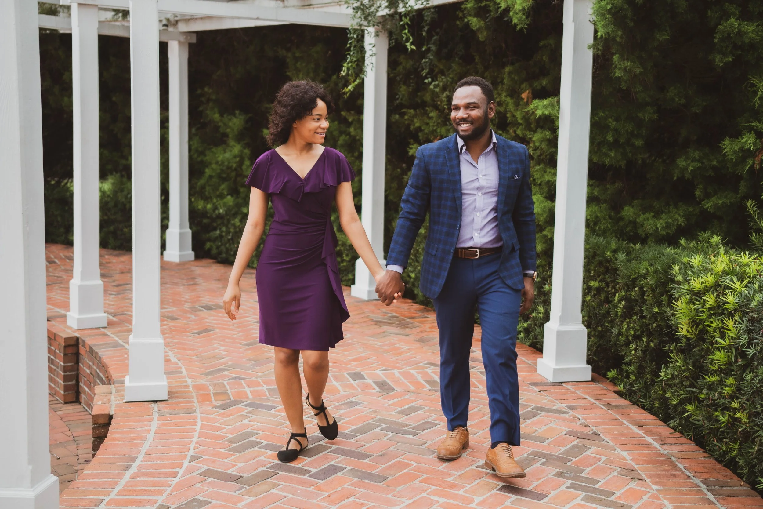 A couple dressed in formal attire walking hand in hand along a brick pathway with greenery on both sides.