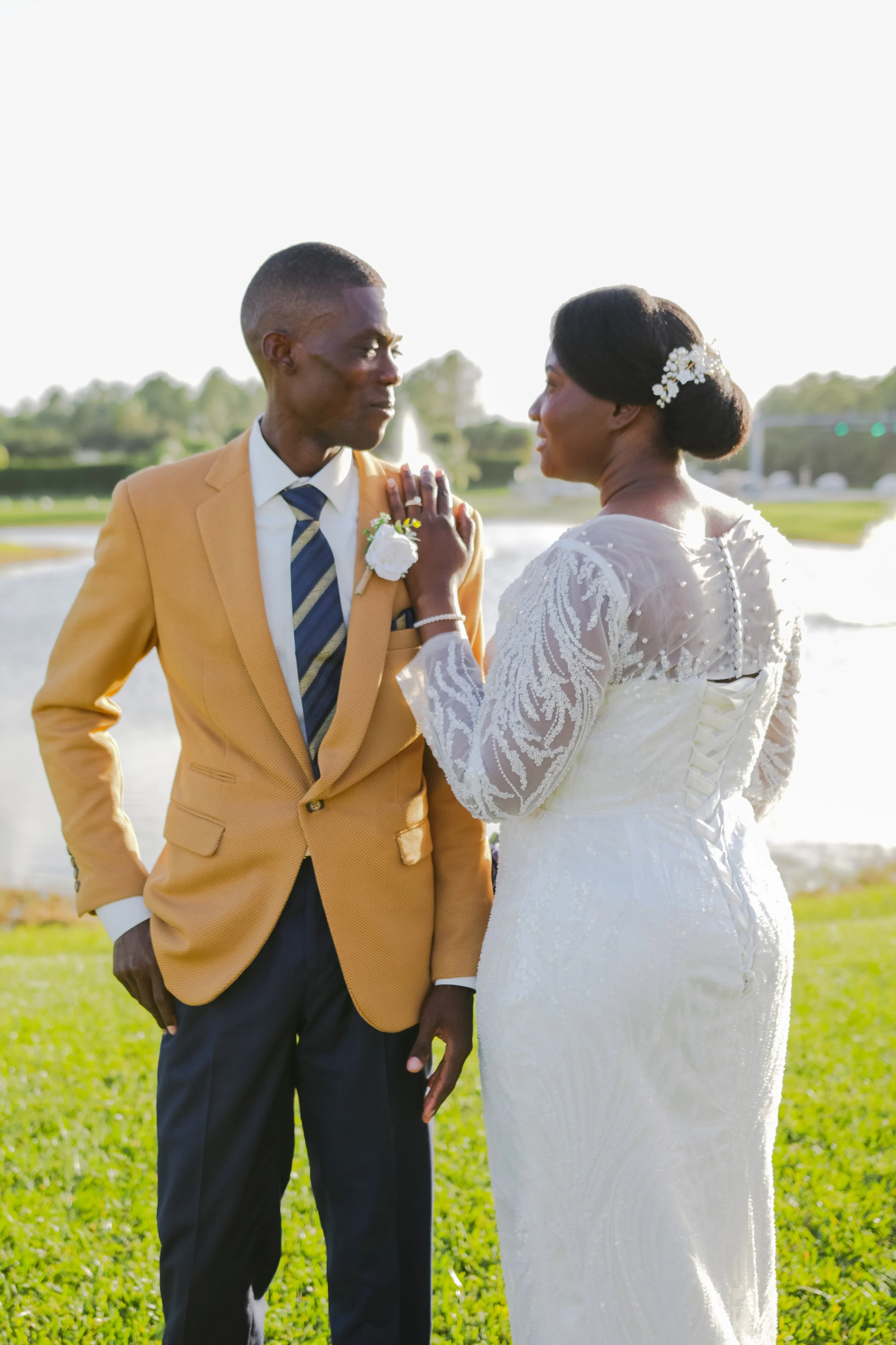 A bride and groom standing outdoors near a lake, dressed in wedding attire, sharing a tender moment.