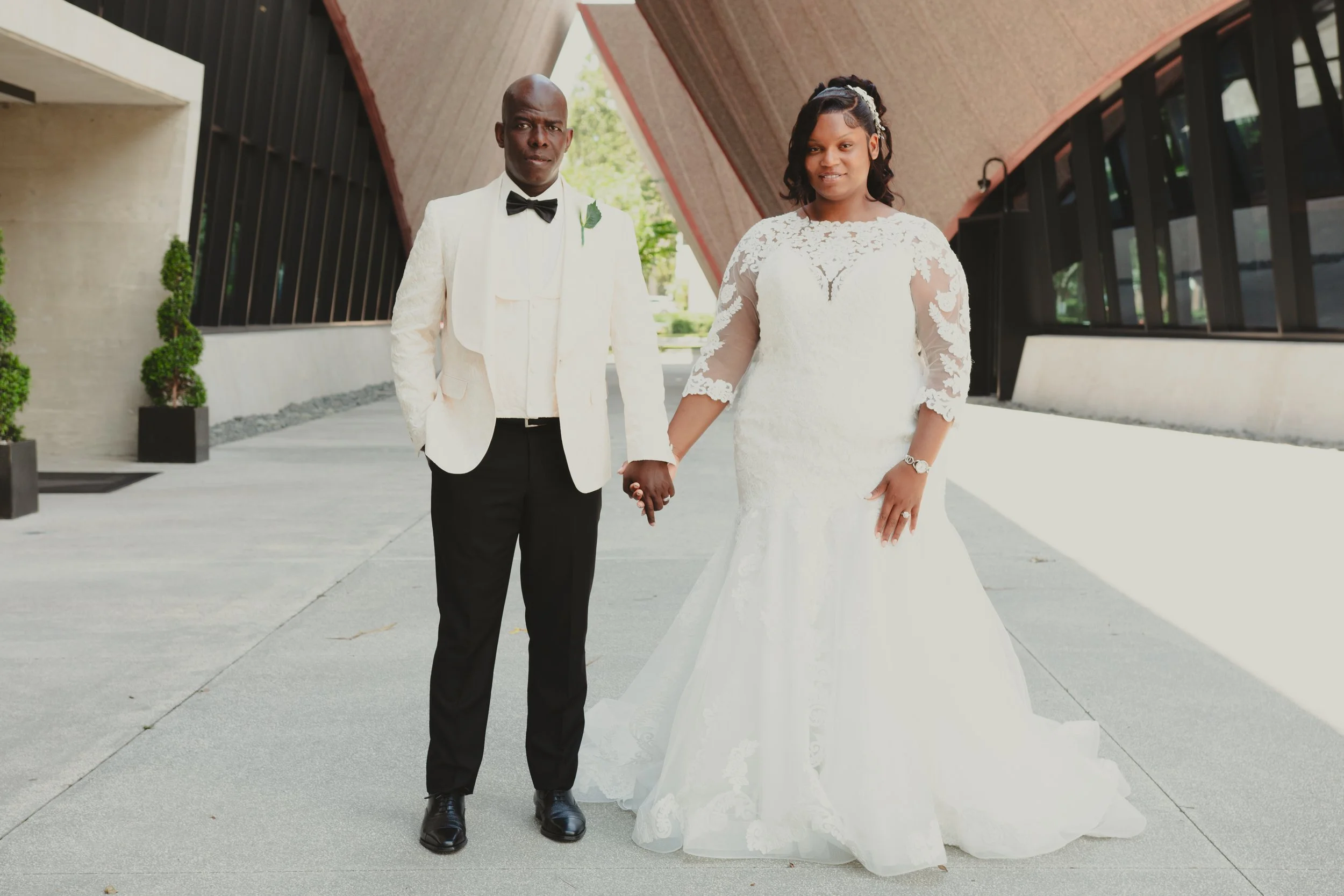 A newlywed couple standing outdoors, holding hands. The groom is wearing a white tuxedo jacket, black pants, and a black bow tie. The bride is in a white lace wedding gown with sheer lace sleeves, and has a wristwatch on her left wrist.