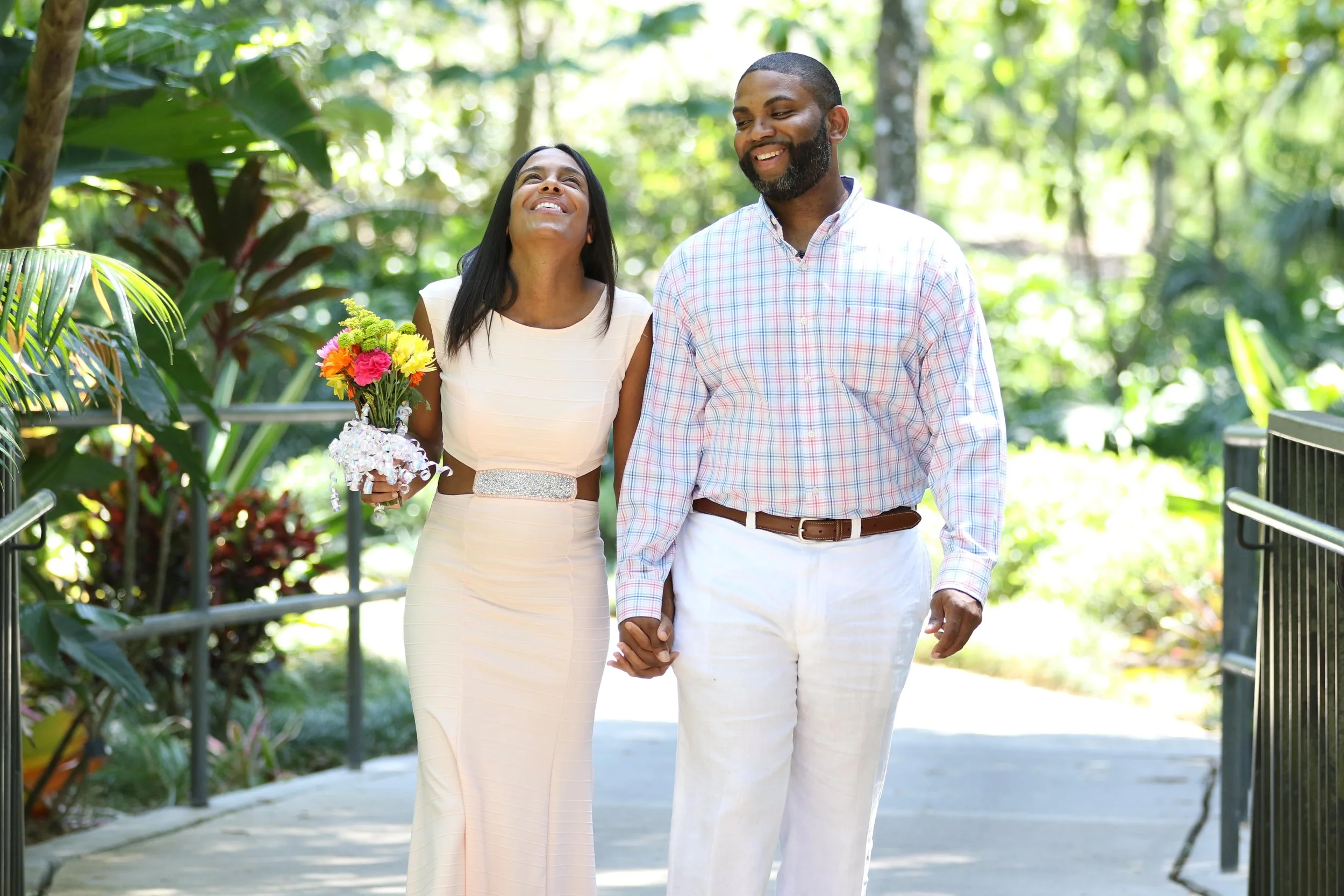 A smiling couple holding hands and walking outdoors in a lush, green garden with sunlight filtering through trees. The woman is holding a bouquet of flowers and wearing a white dress, while the man is dressed in a light-colored, checkered shirt and w