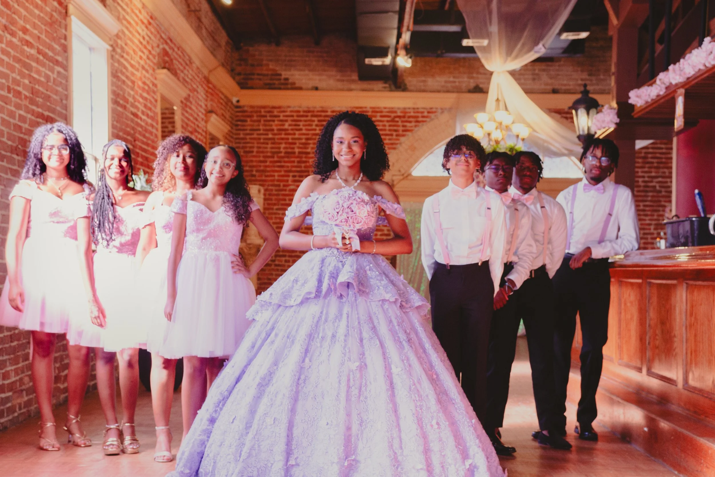 A bride in a lavender ball gown holding a bouquet, standing with six children dressed in formal attire in a decorated brick venue.
