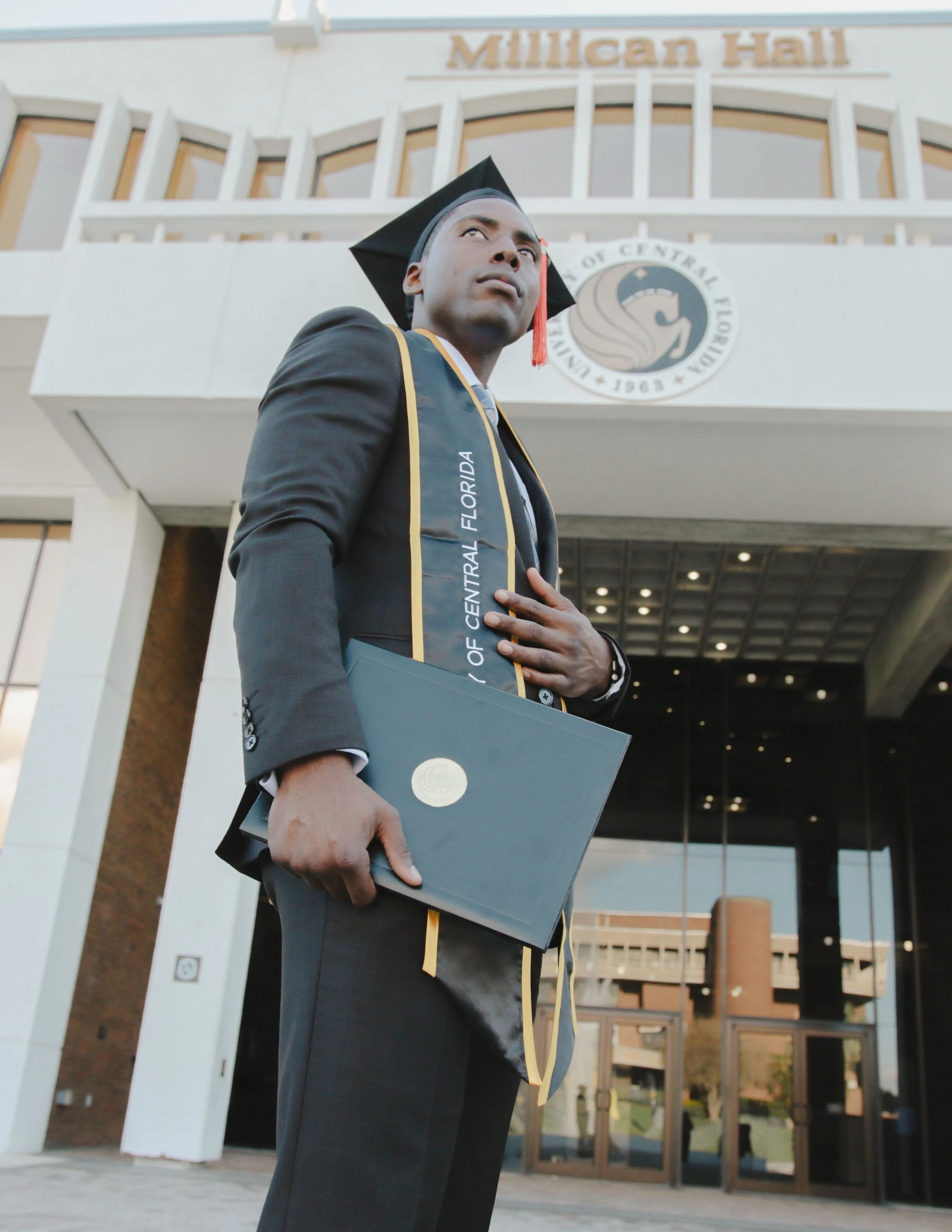 A man in academic regalia standing outside Milligan Hall at the University of Central Florida, holding a diploma with a contemplative expression.