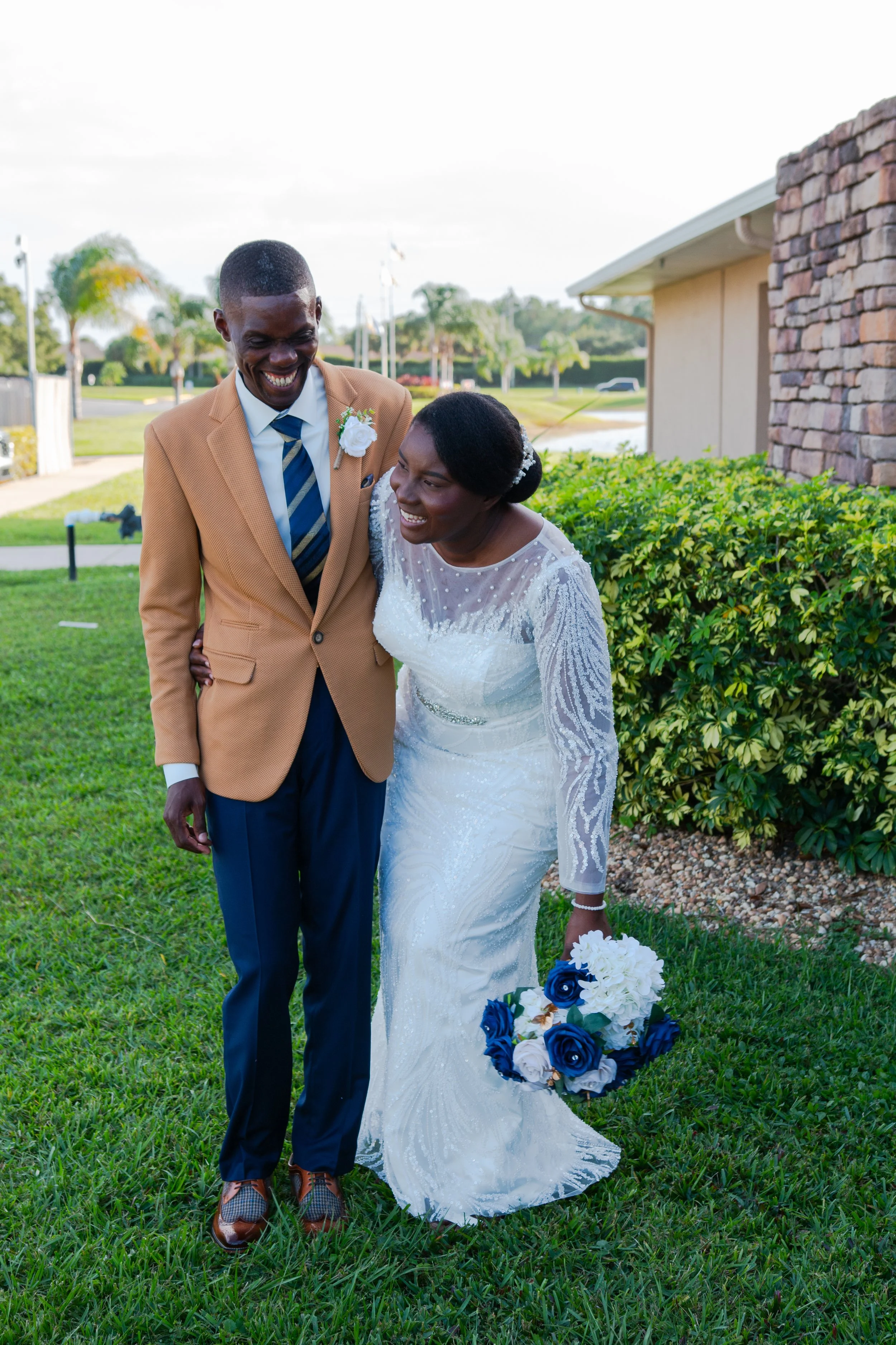 A bride and groom walking together outdoors, smiling, with the bride holding a bouquet of blue and white flowers.