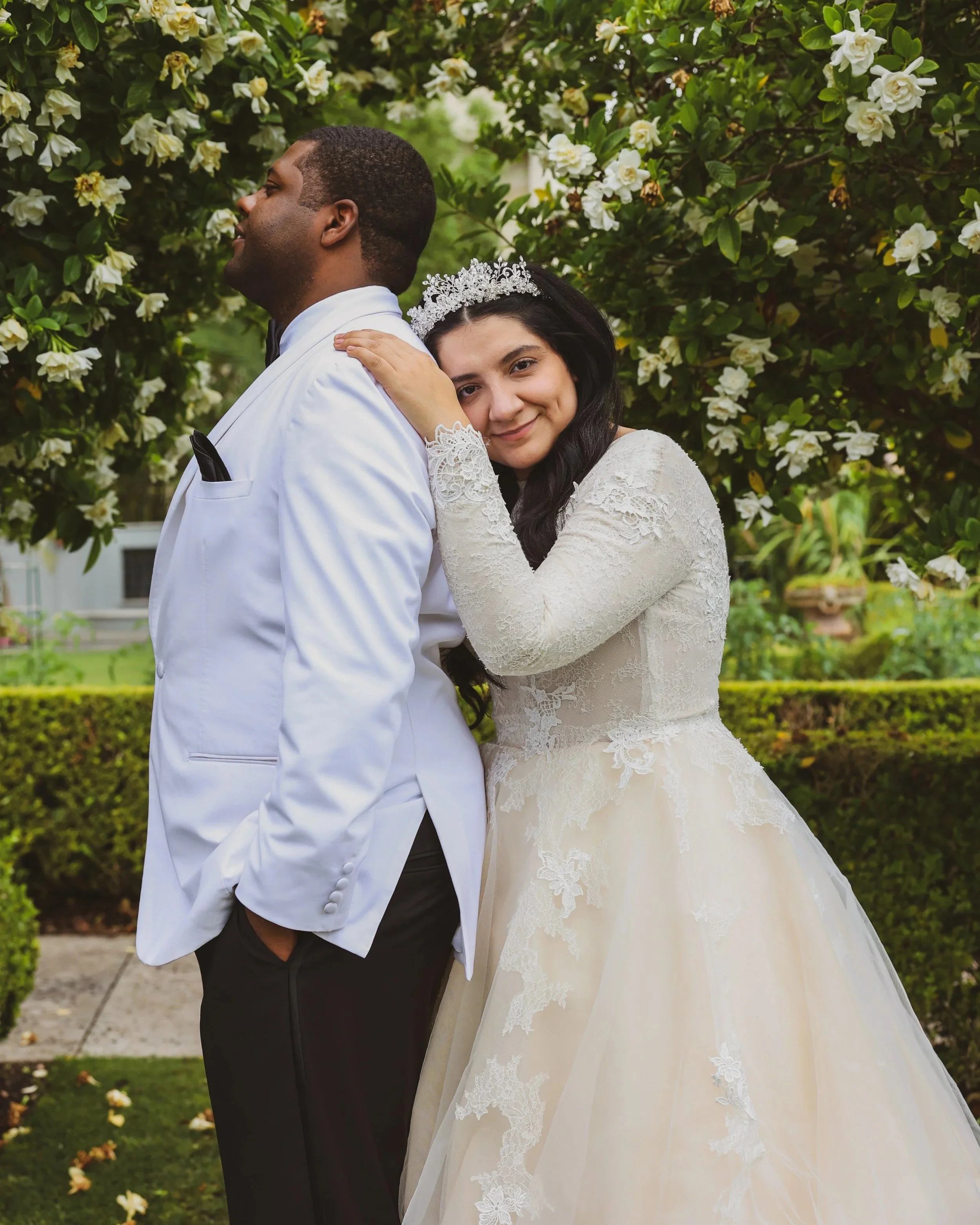 A bride in a lace wedding gown and tiara leaning on a groom in a white tuxedo, posing outdoors near blooming white flowers and greenery.