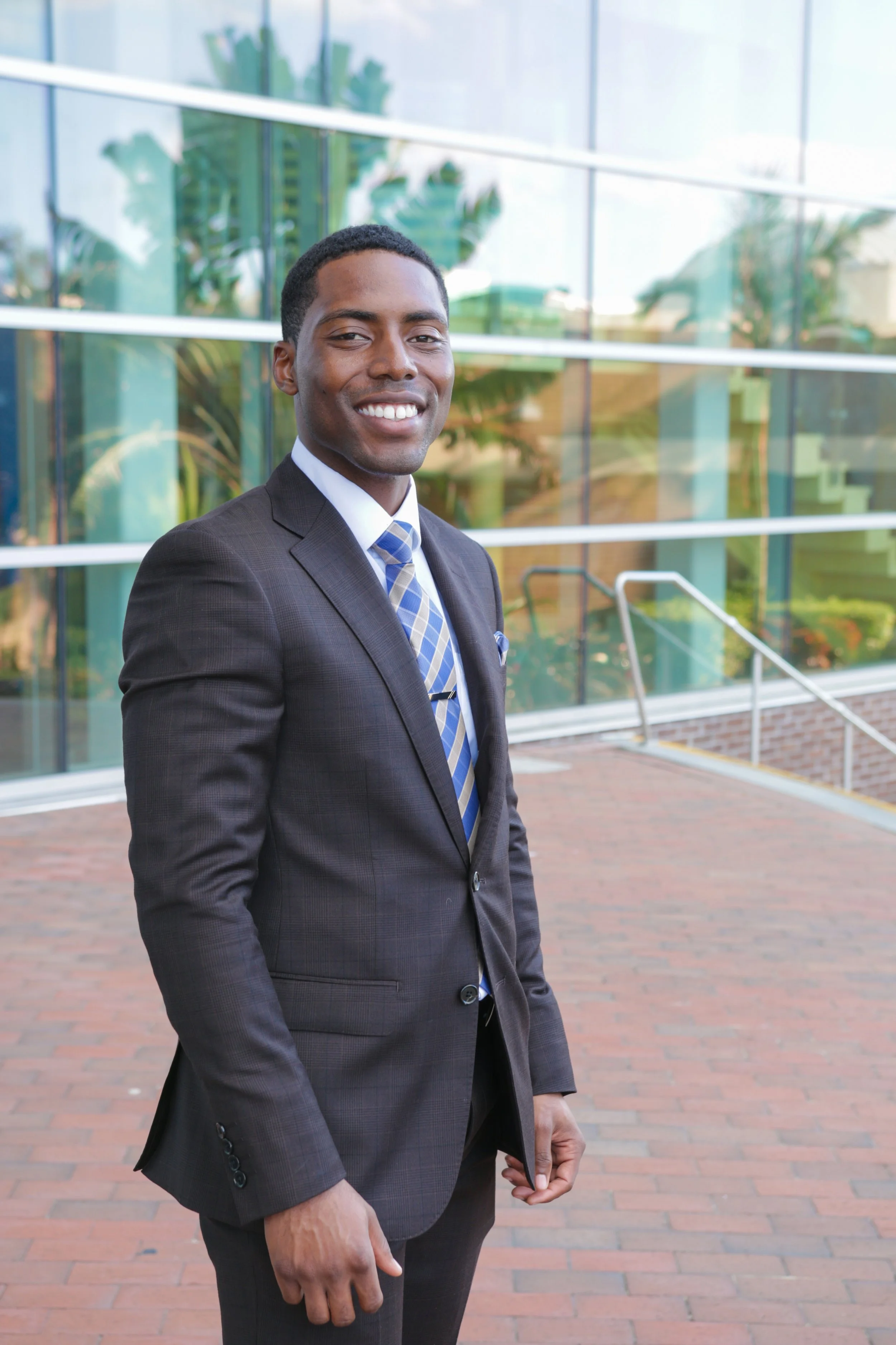 A smiling man dressed in a dark suit, white shirt, and striped blue tie, standing outside a modern glass building with greenery reflected in the windows.