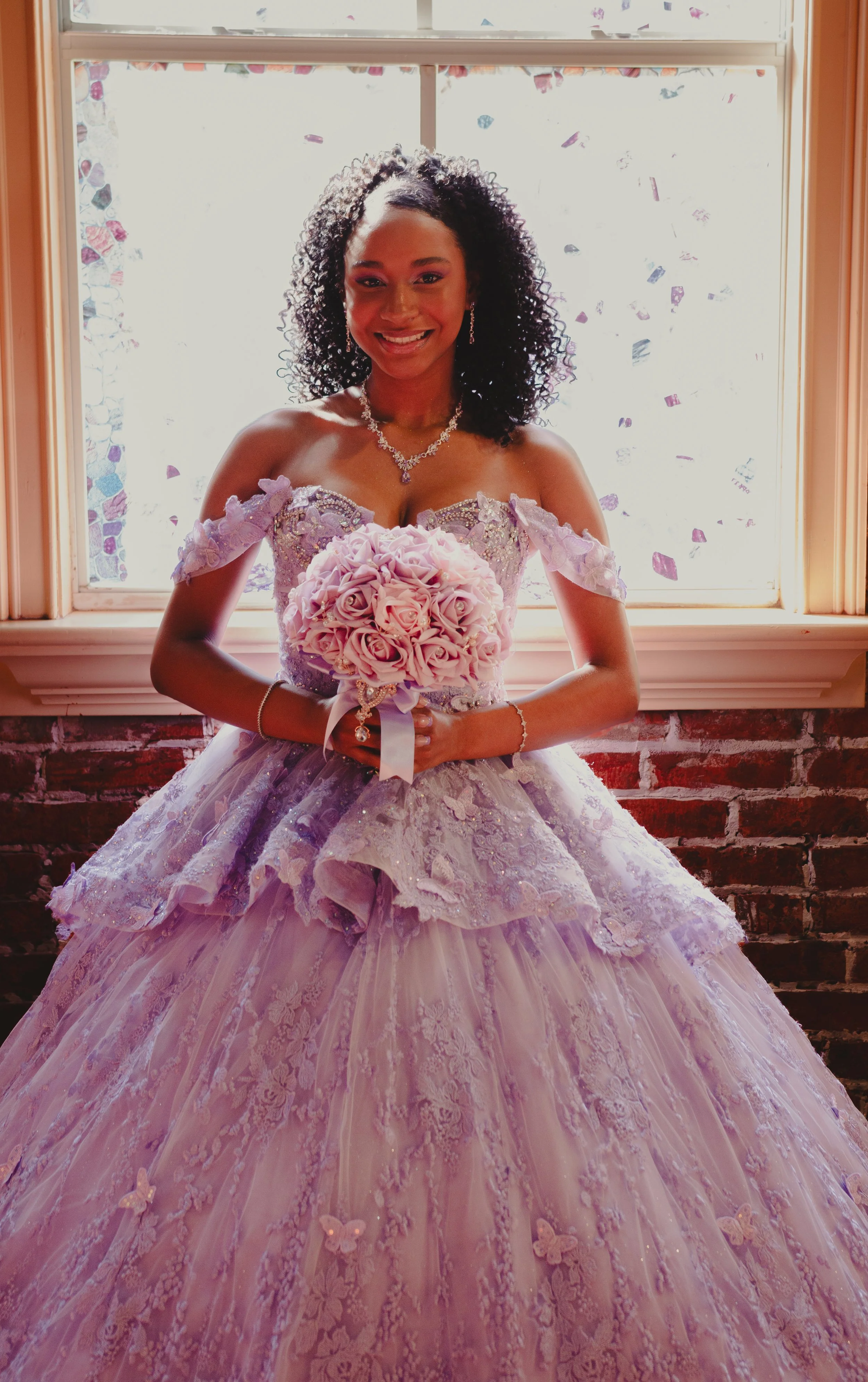 A smiling woman in a lavender wedding gown holding a bouquet of light pink roses, standing in front of a window with confetti.
