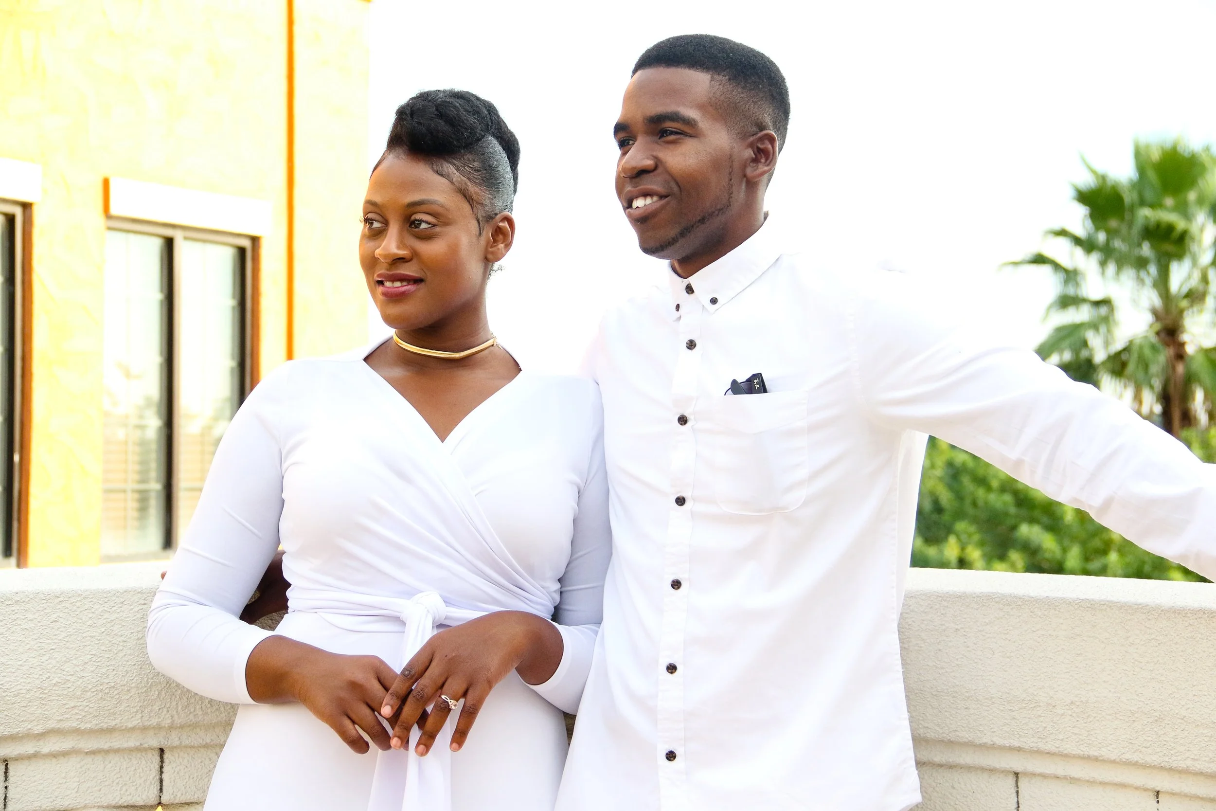 A man and woman standing together outdoors during daytime, dressed in white, with the man smiling and the woman looking thoughtful. The woman has short black hair styled in an updo, and the man has a short haircut. They are standing near a cream-colo