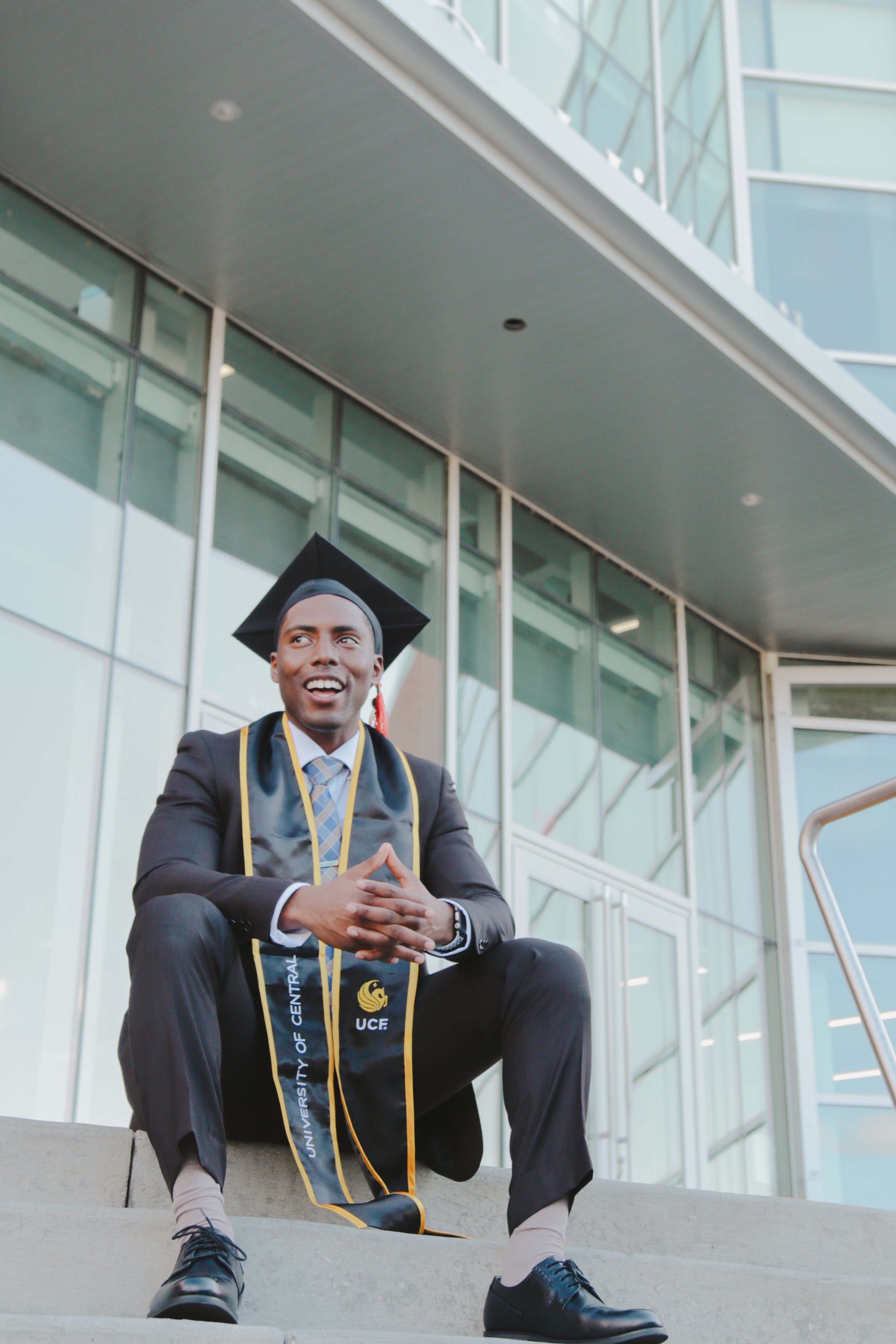 A young man in graduation cap and gown sitting on steps outside a modern glass building, smiling, with university stole and medal.