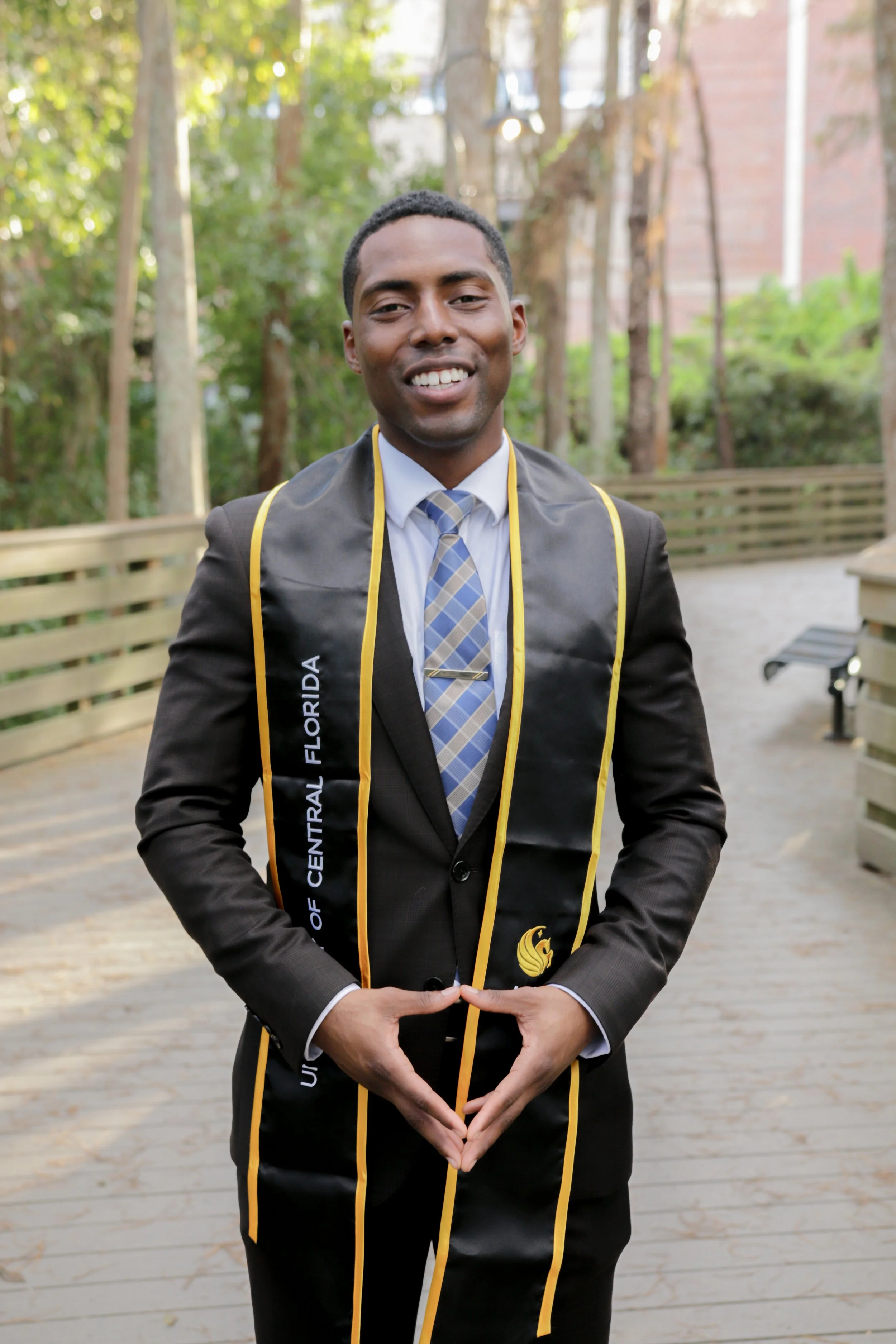 A young man wearing a business suit and tie, smiling, with a graduation stole that says 'University of Central Florida, Florida.' He stands outdoors on a wooden pathway surrounded by trees.