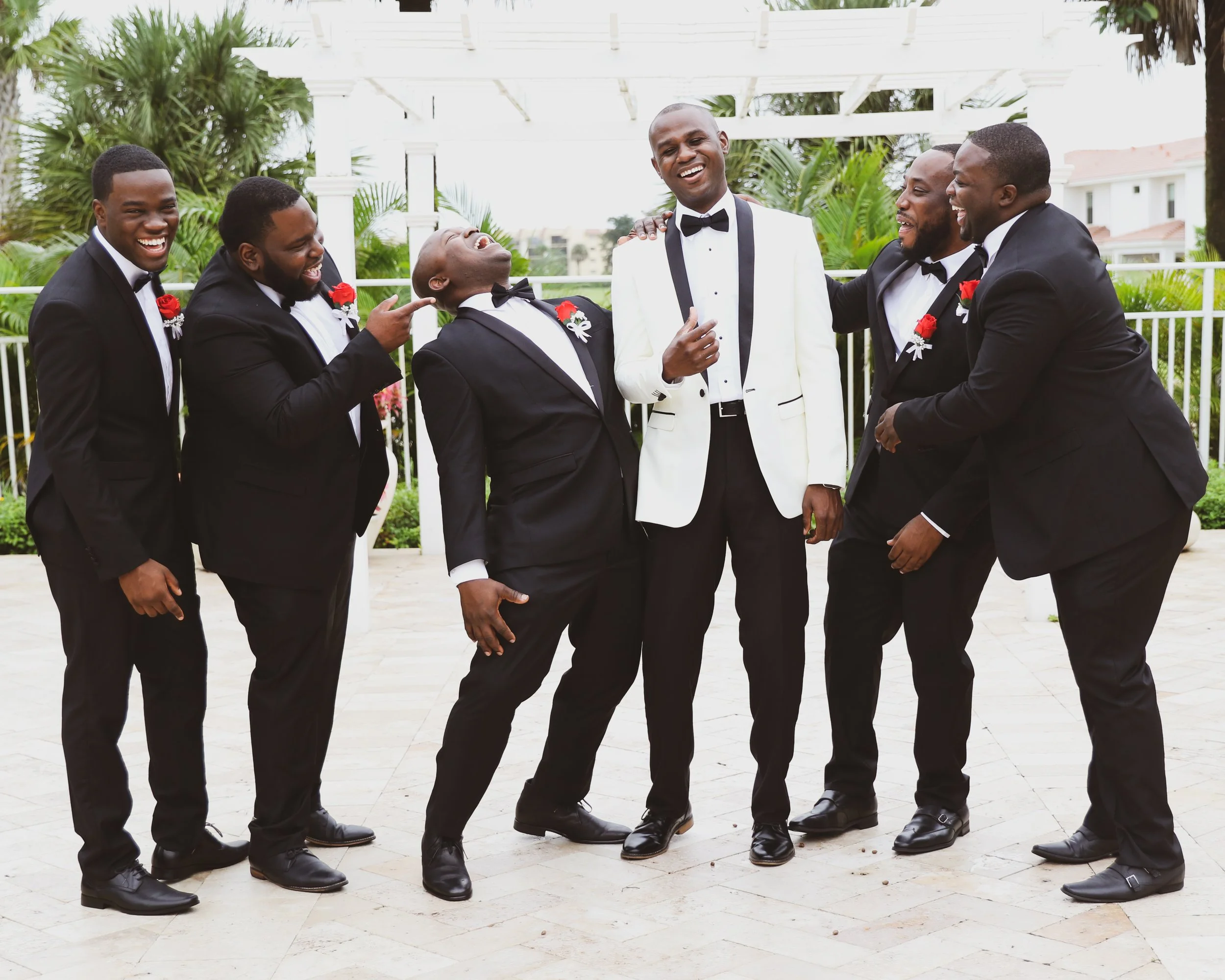 A group of seven men dressed in tuxedos, with the man in the middle wearing a white tuxedo jacket, celebrating outdoors with palm trees and a white gazebo in the background.