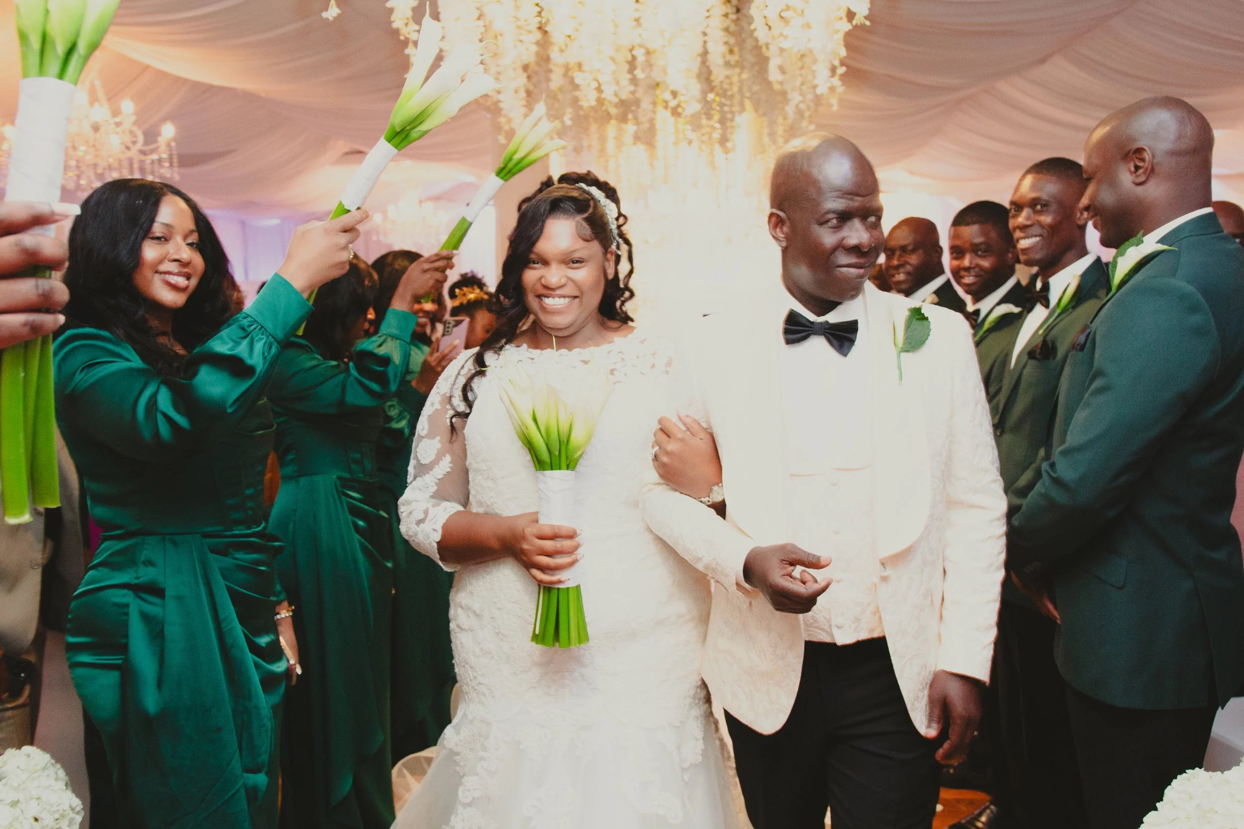 Wedding couple walking down the aisle while friends and family hold white calla lilies in the air, with a decorated ceiling and chandelier in the background.
