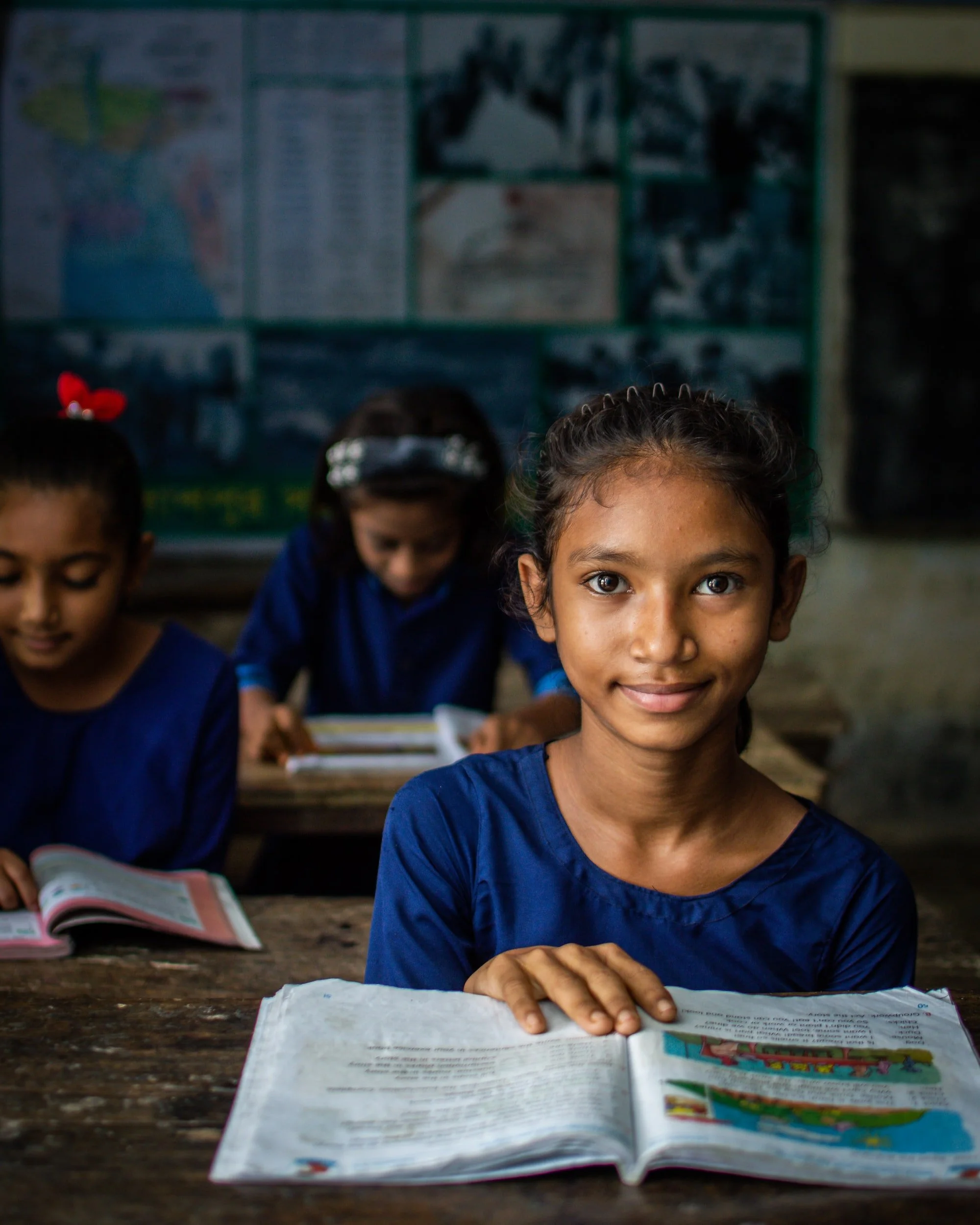 A young girl with dark hair and brown skin smiling at the camera in a classroom, sitting at a wooden desk with open textbooks, with other girls reading in the background.