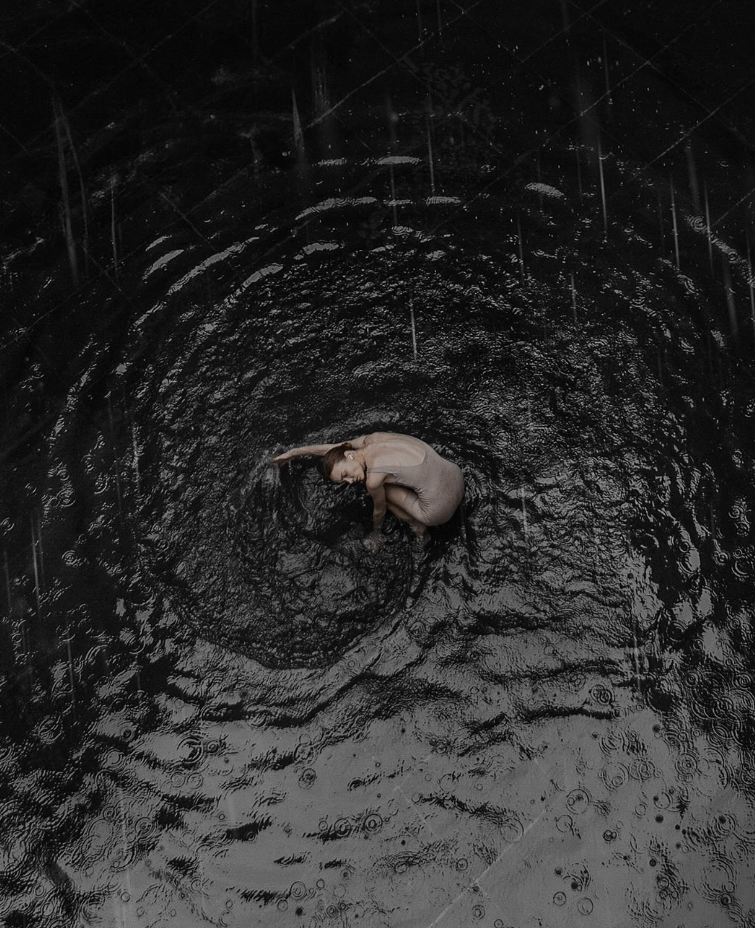 A woman in a beige dress crouches in the center of a dark, reflective, rain-covered surface, creating circular ripples around her, with raindrops falling all around.