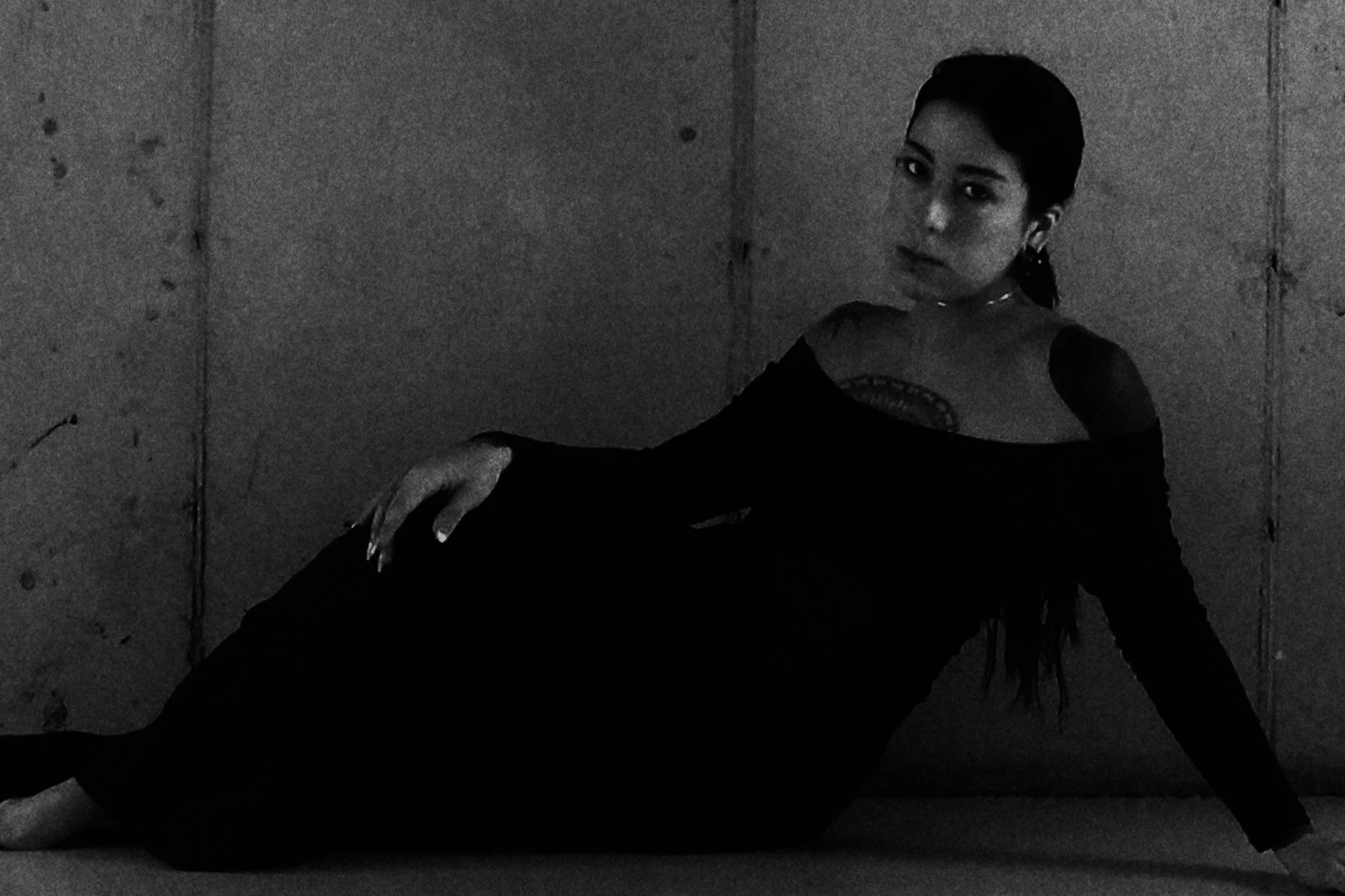 A woman wearing a black off-the-shoulder dress sitting against a textured wall, looking at the camera.