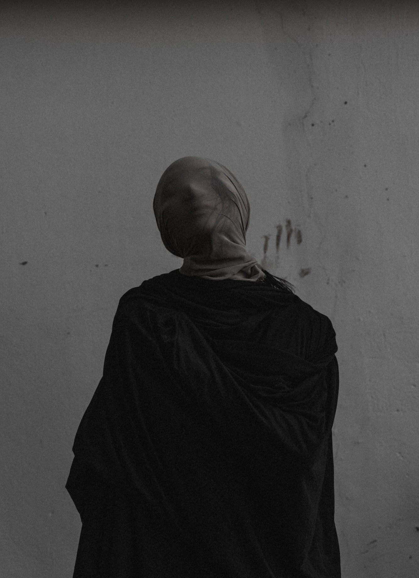 A person wearing a beige head covering and black clothing standing with their back to the camera against a textured gray wall.