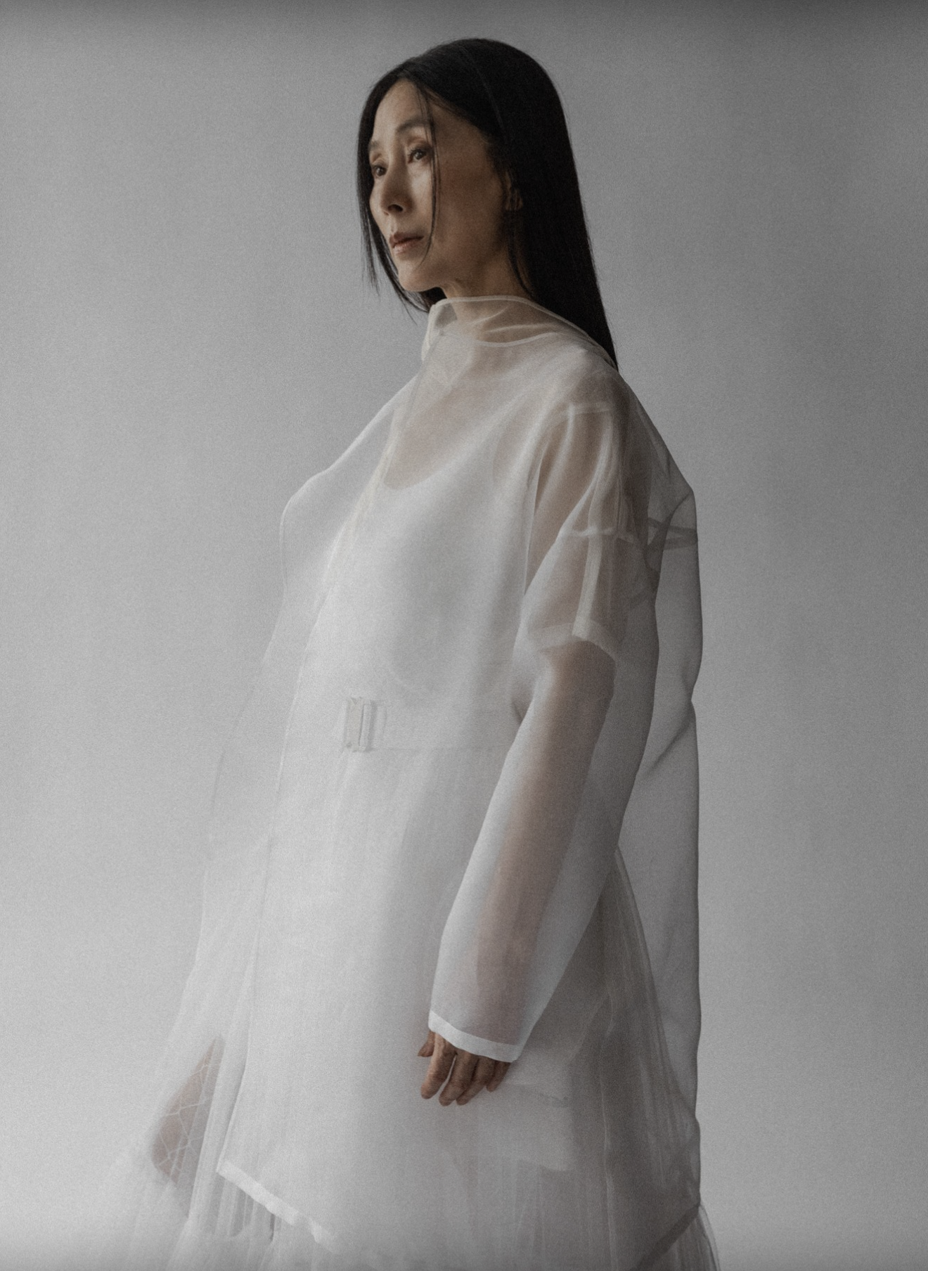 A woman with long dark hair wearing a sheer, white, layered dress stands against a plain gray background, looking to her right with a contemplative expression.
