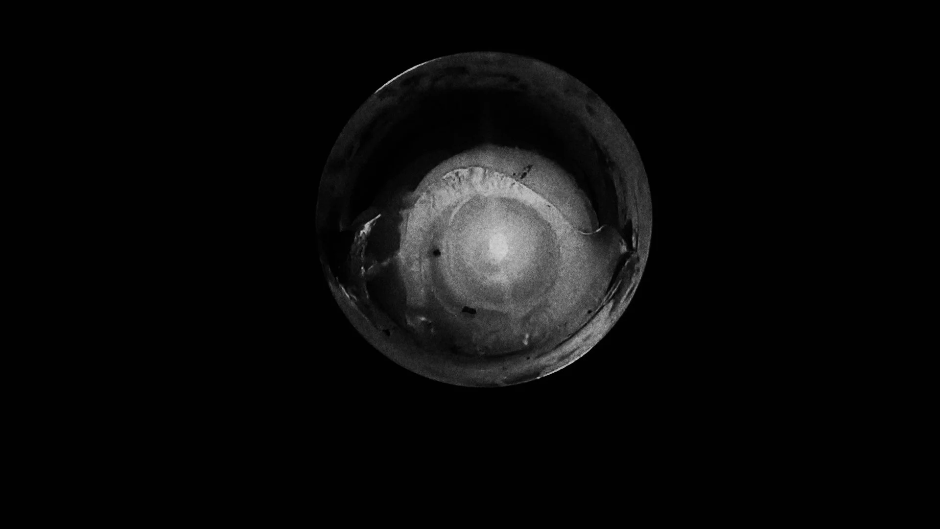 An overhead view of a metallic, circular object with a luminous, textured surface, captured in black and white against a dark background.