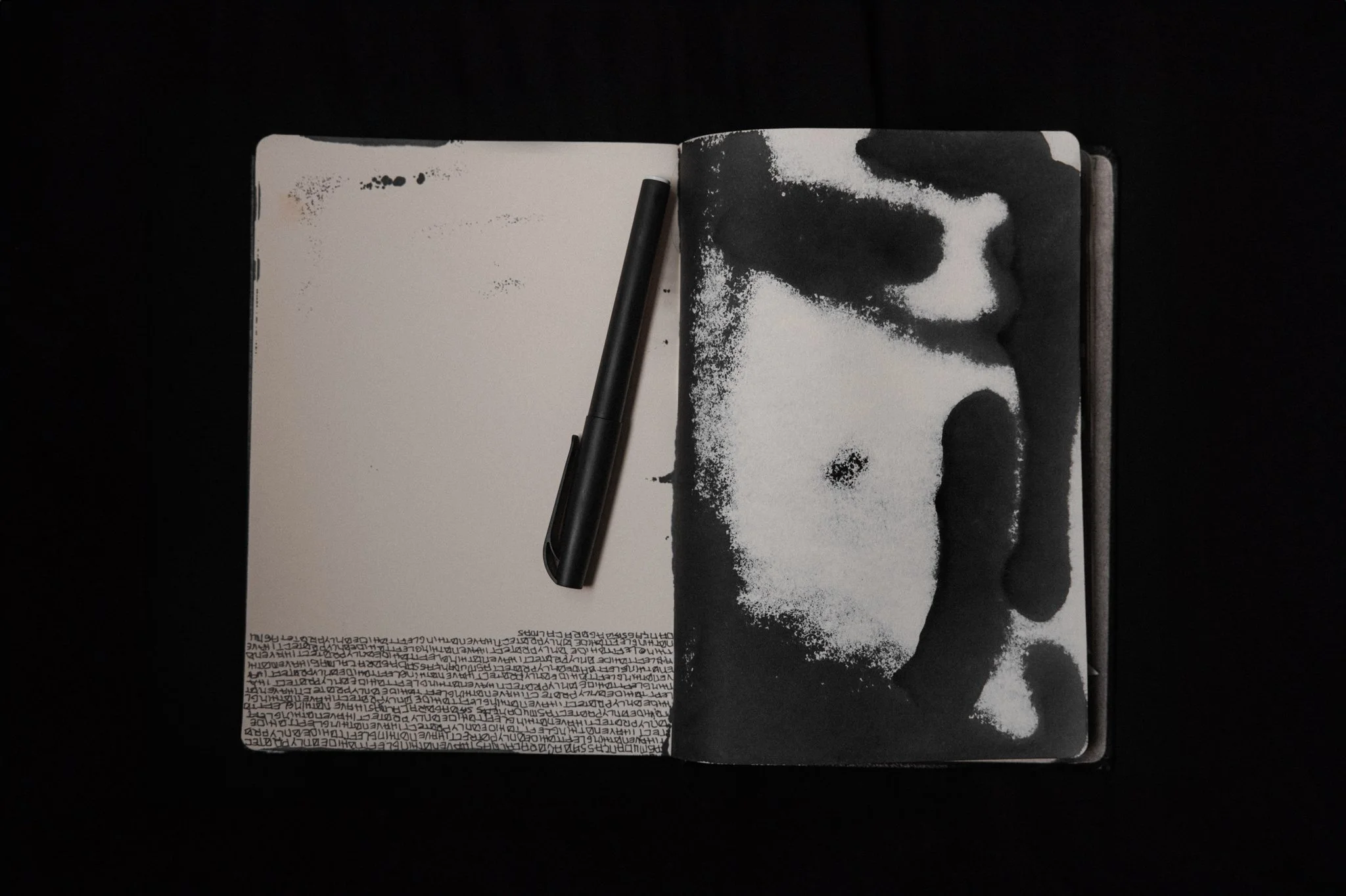 An open sketchbook with a black marker resting on the left page, and a portrait of a person's face in black and white ink on the right page, set against a dark background.