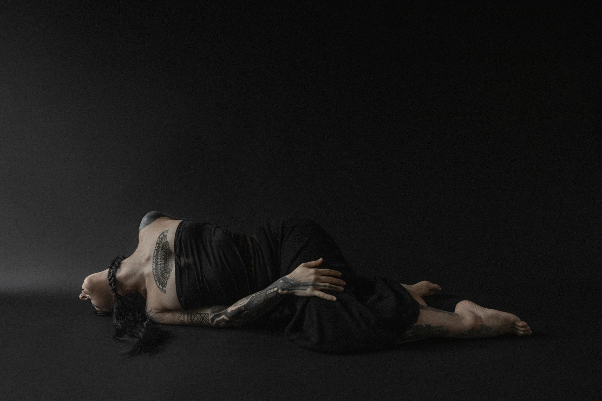 A woman lying on her side on a black surface with a black background, wearing a black dress, with tattoos on her arms and chest, and black hair.