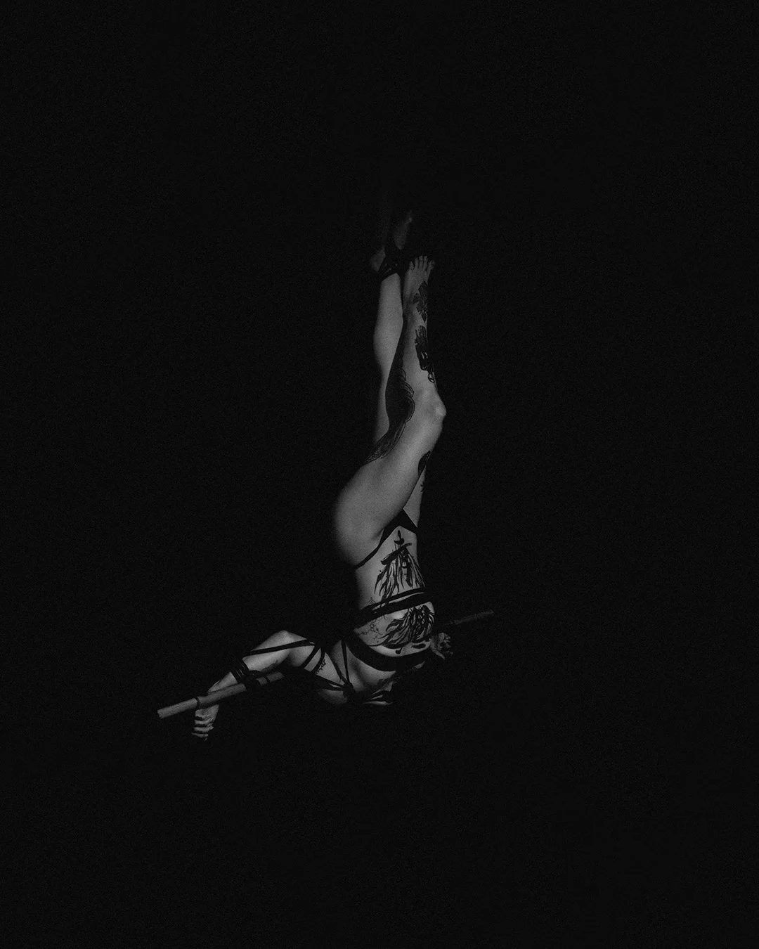 A person with tattoos in a harness, performing a yoga pose on a pole in a dark setting with minimal lighting.