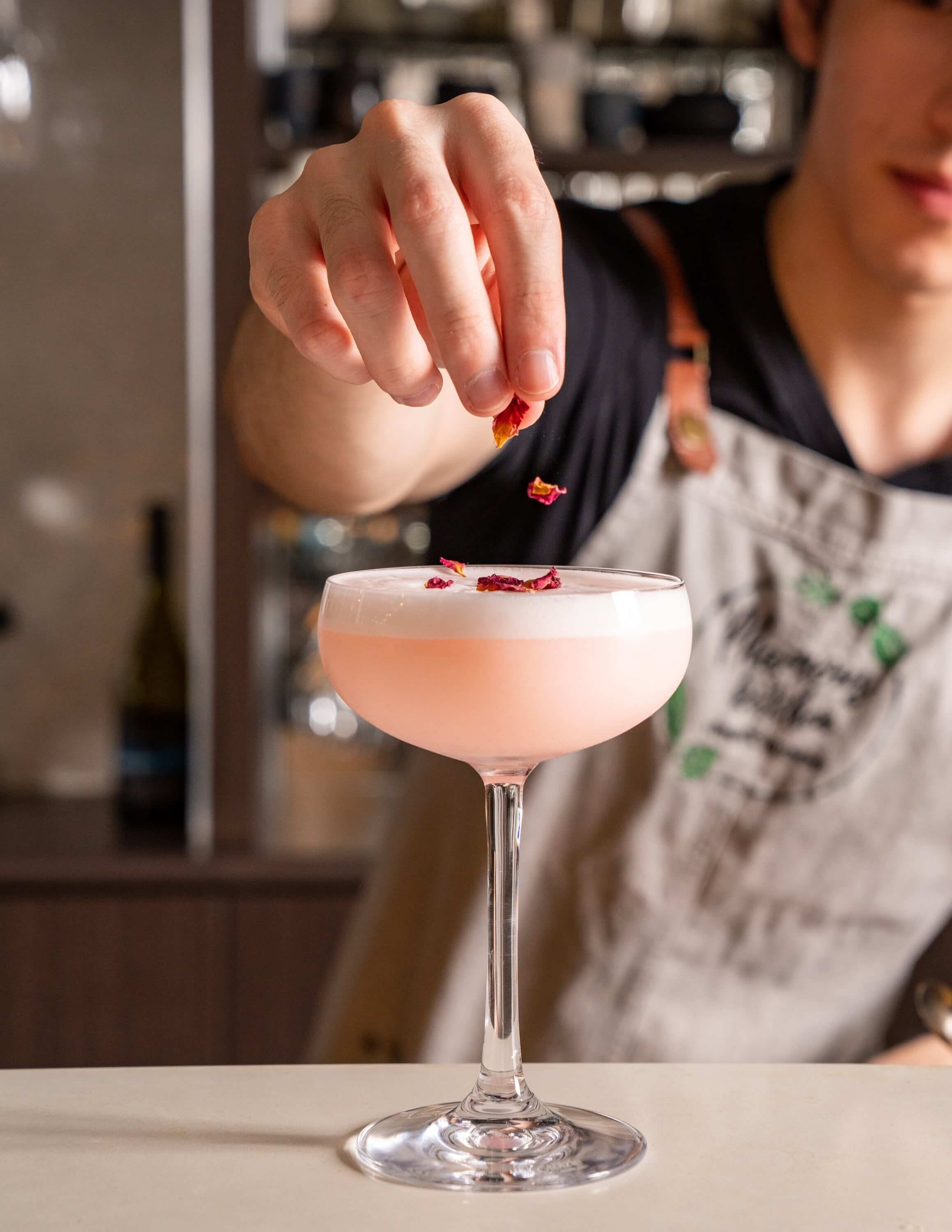  bartender adding dried rose petles to the top of a pink cocktail 