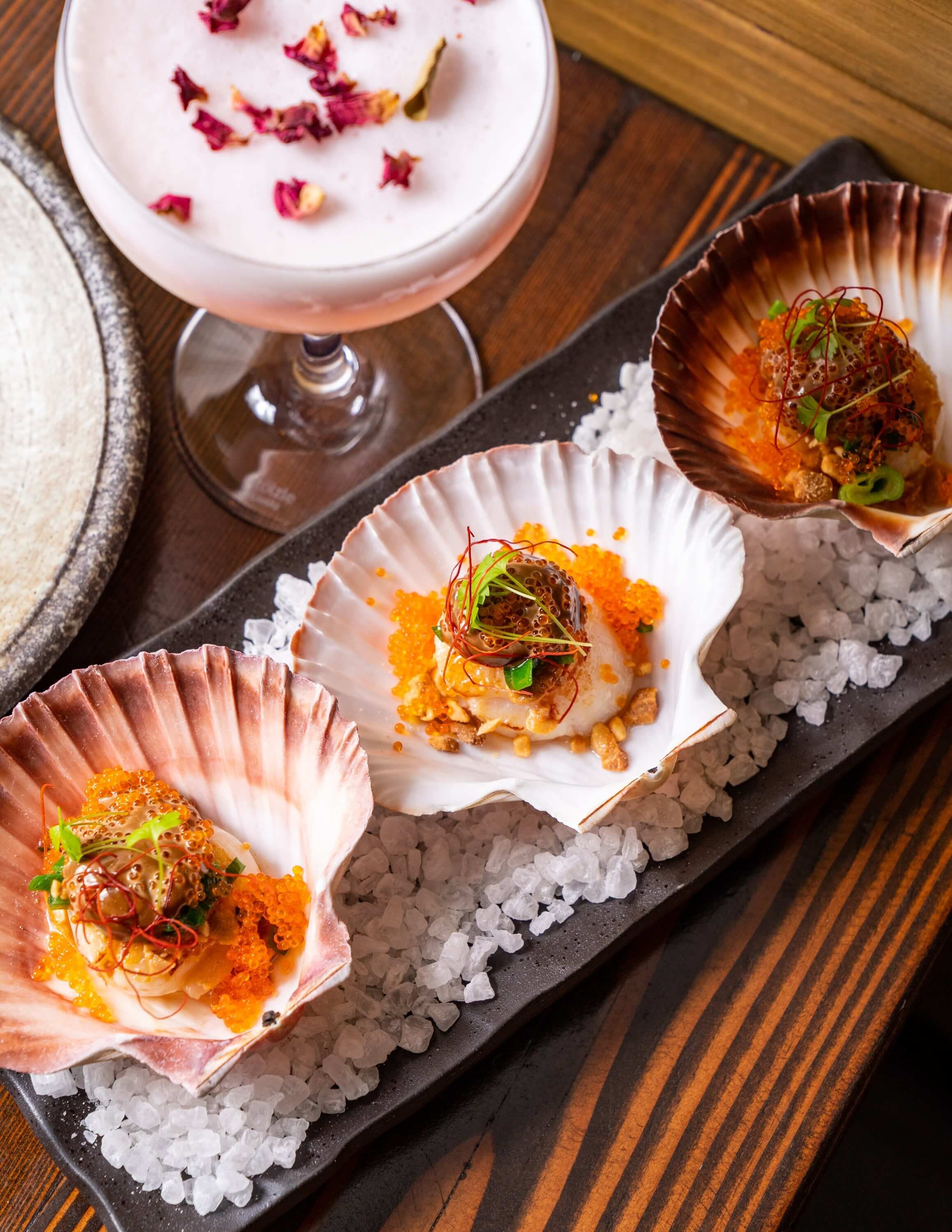  Street-style Seared Scallop served with black garlic aioli, topped with shallot oil, peanut, tobiko and herbs on a dish with rock salt and a rose cocktail 