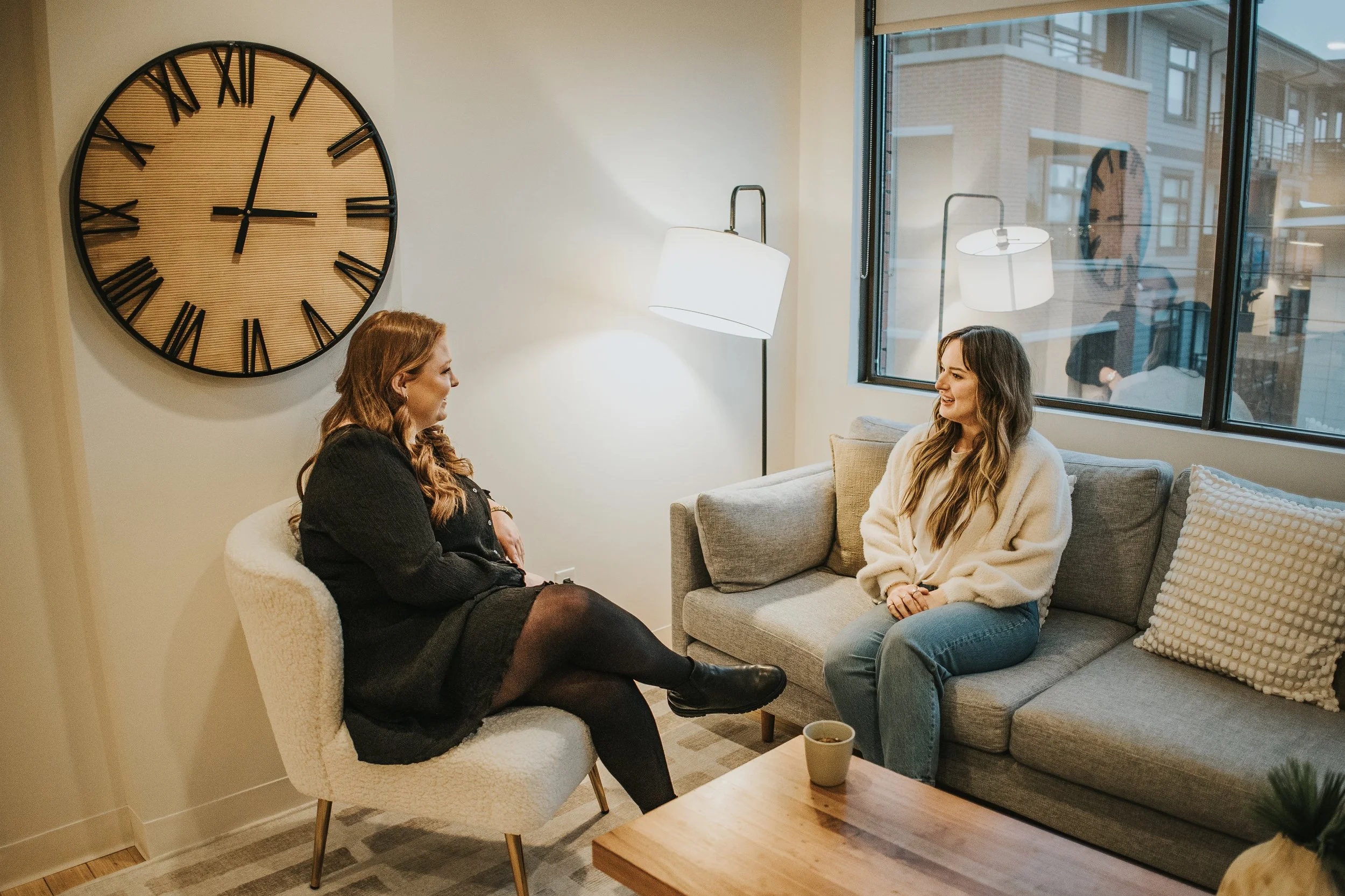 Two women sitting and talking in a cozy living room, one on a white armchair and one on a gray sofa, with large wall clock and a window showing apartment buildings outside.