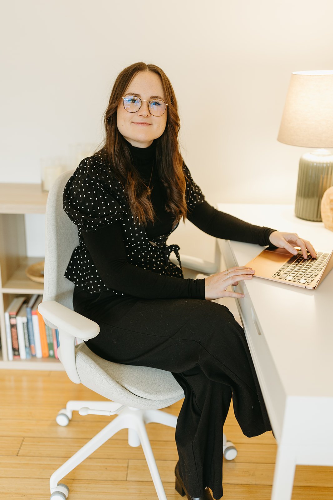 A woman with brown hair, glasses, and wearing a black polka dot top and black pants, sitting on a white office chair at a desk with a rose gold laptop, working inside a modern, well-lit office.