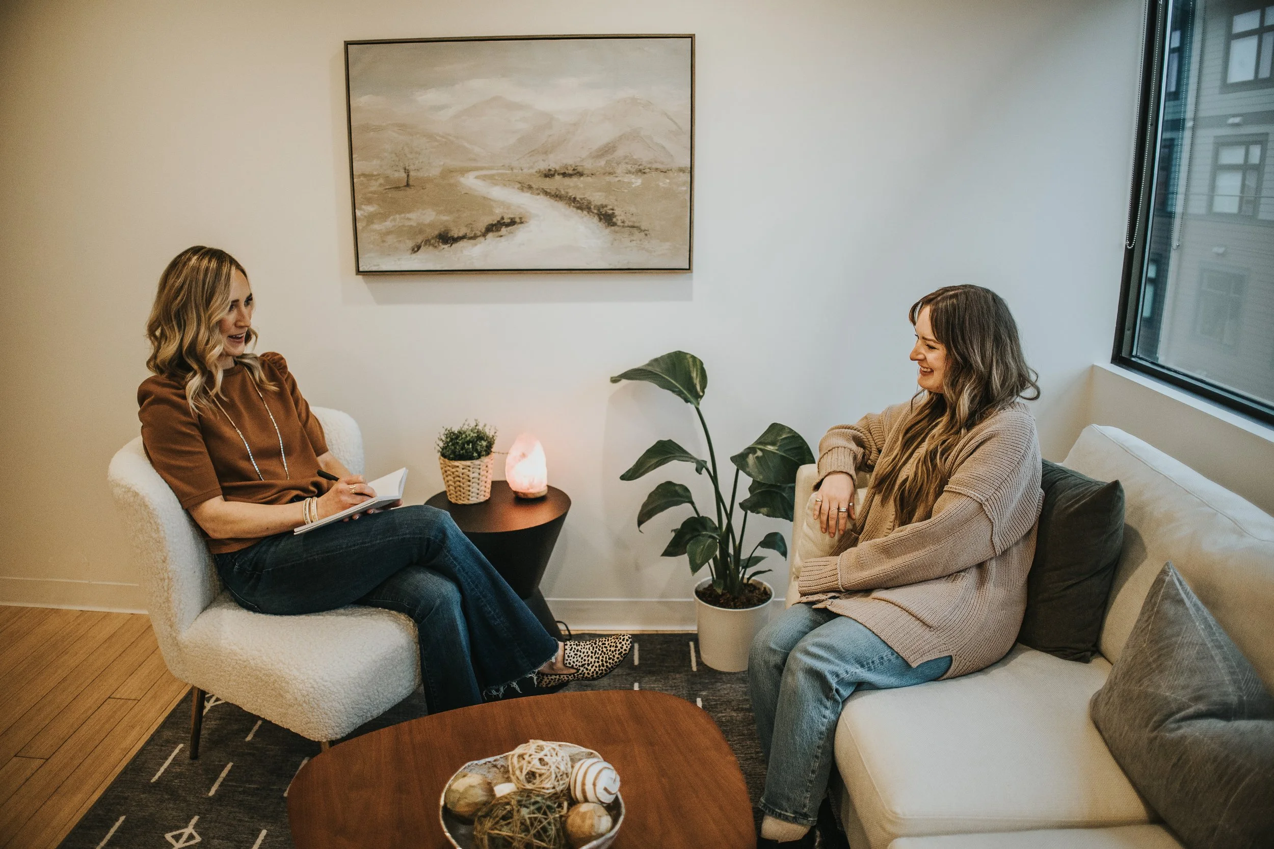 Two women sitting in a cozy living room, having a conversation. One woman is taking notes, while the other is smiling. The room has a plant, a painting on the wall, a side table with a salt lamp, and a sofa with cushions.