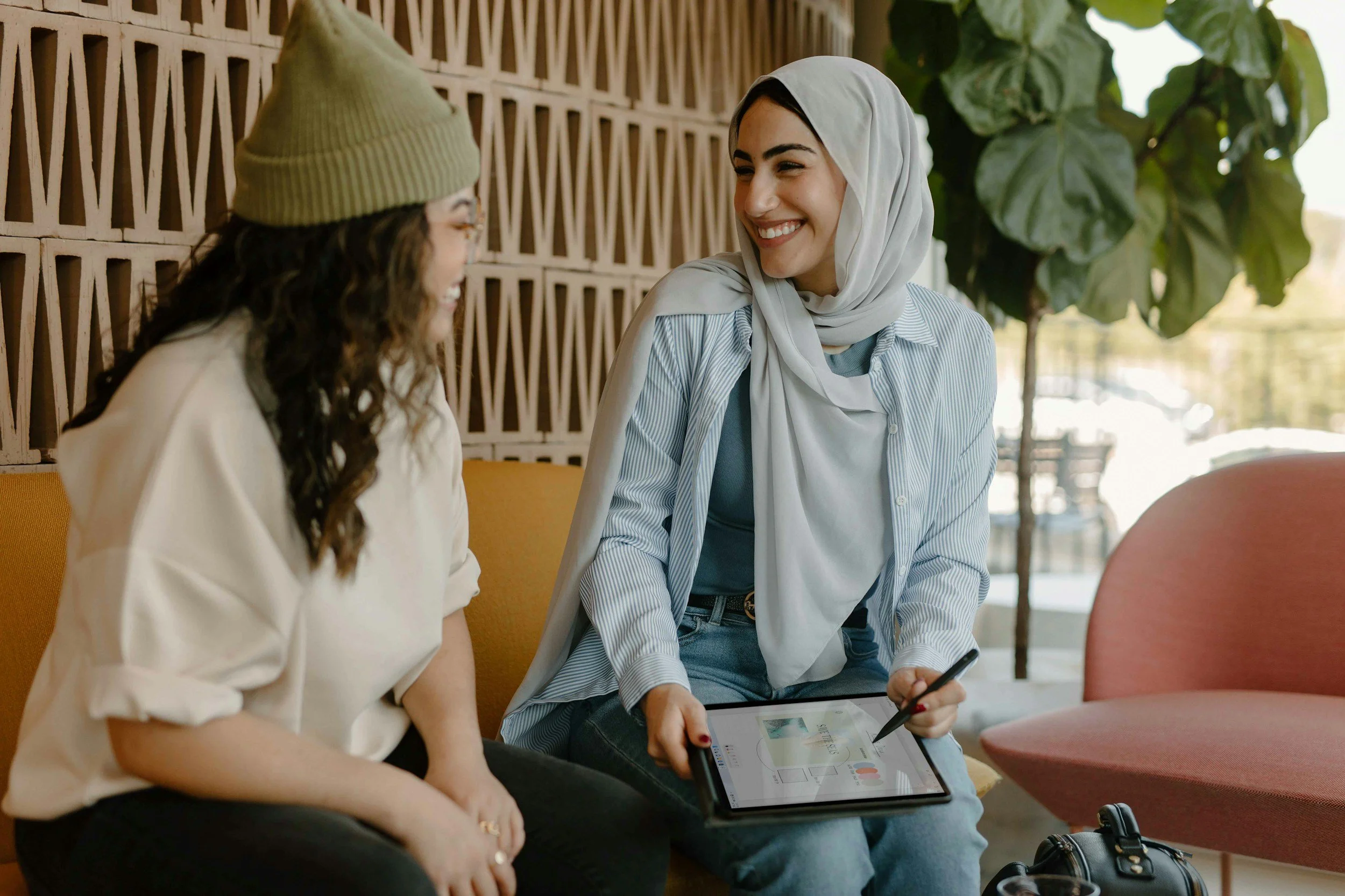 Two women sitting on a yellow couch, engaged in conversation and smiling. One woman holds a digital tablet with a stylus. The other woman wearing a beanie and glasses is listening attentively. Bright indoor setting with a decorative wall and large green plant in the background.