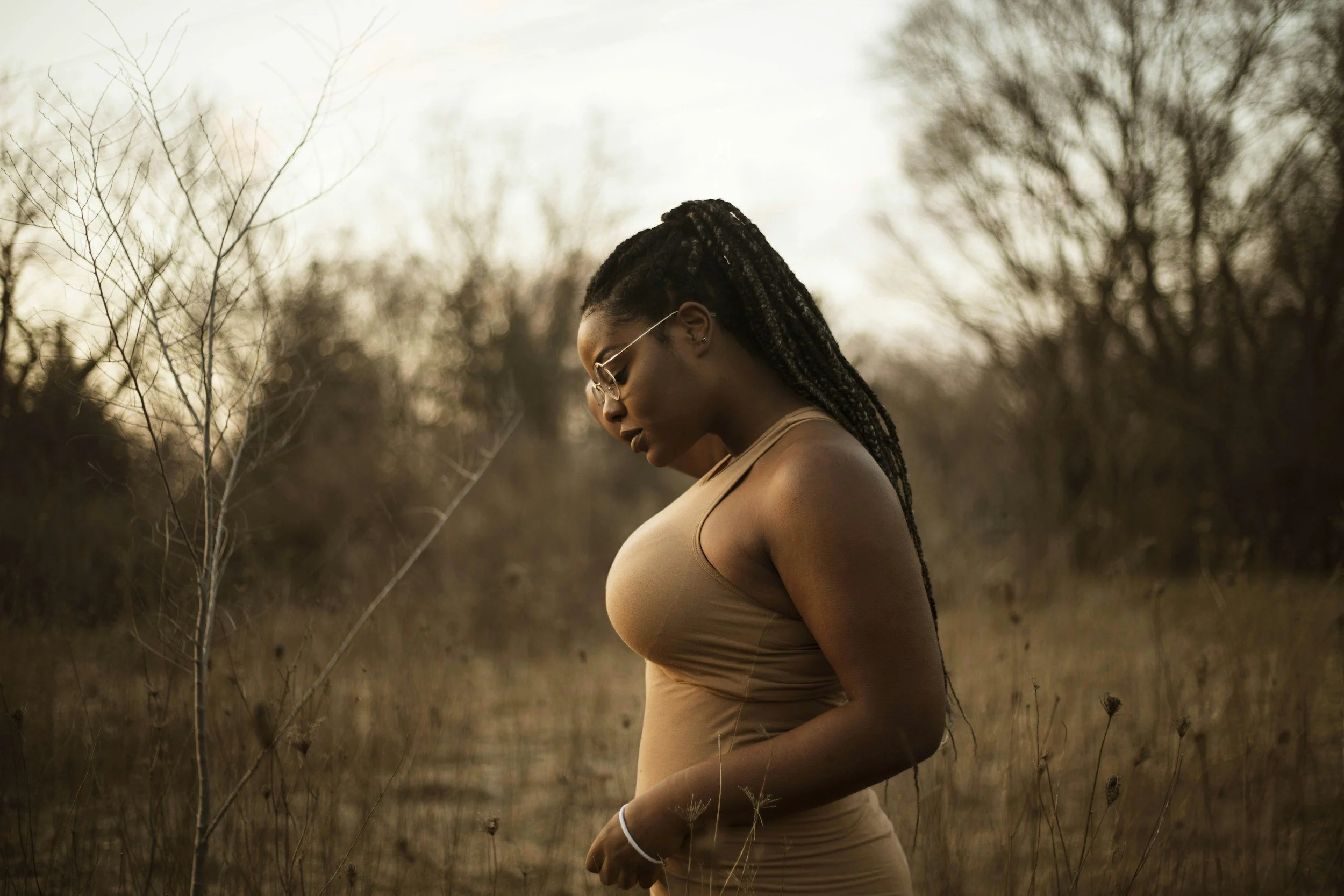 A woman with braided hair and glasses standing in a field of dry grass with bare trees in the background during cloudy weather.