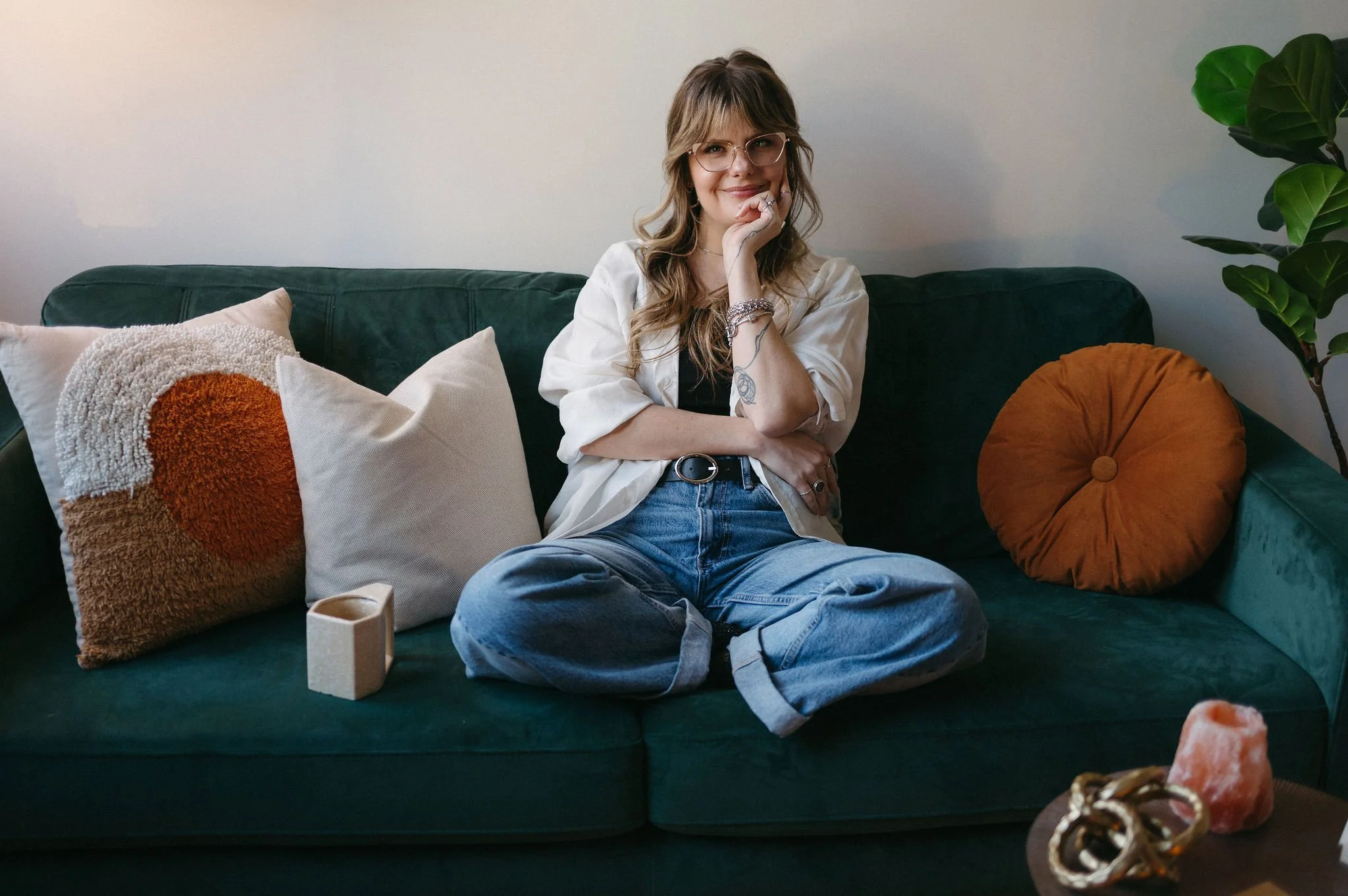 A woman sitting cross-legged on a green velvet sofa, surrounded by decorative pillows, a succulent plant on the right, and items on the coffee table in front of her.