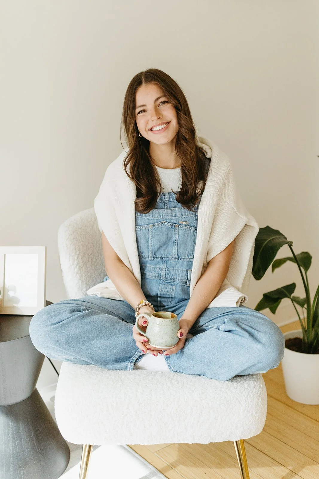 A young woman sitting cross-legged on a white armchair, smiling and holding a mug in her hands. She has long brown hair, is wearing a white t-shirt, denim overalls, and a light sweater draped over her shoulders. There is a potted plant behind her and a framed picture on a small table to her left.