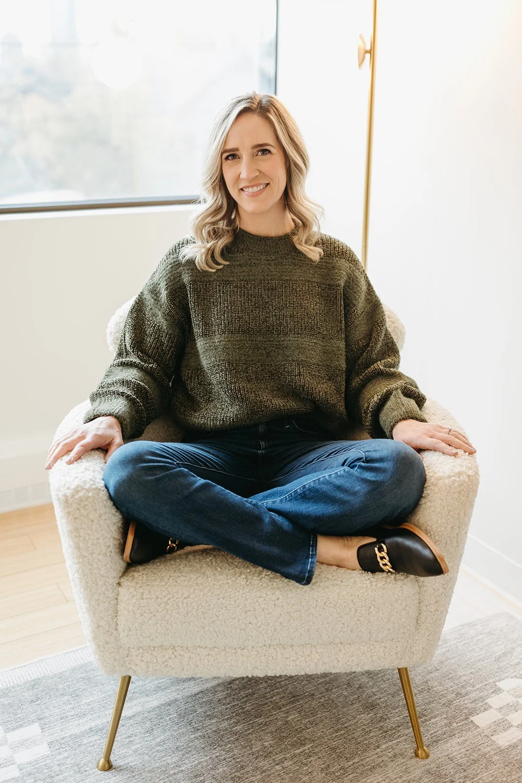 Woman sitting cross-legged on a cozy, cream-colored armchair with gold legs, in front of a large window, wearing a dark green sweater, blue jeans, and black shoes with gold chain details, smiling.