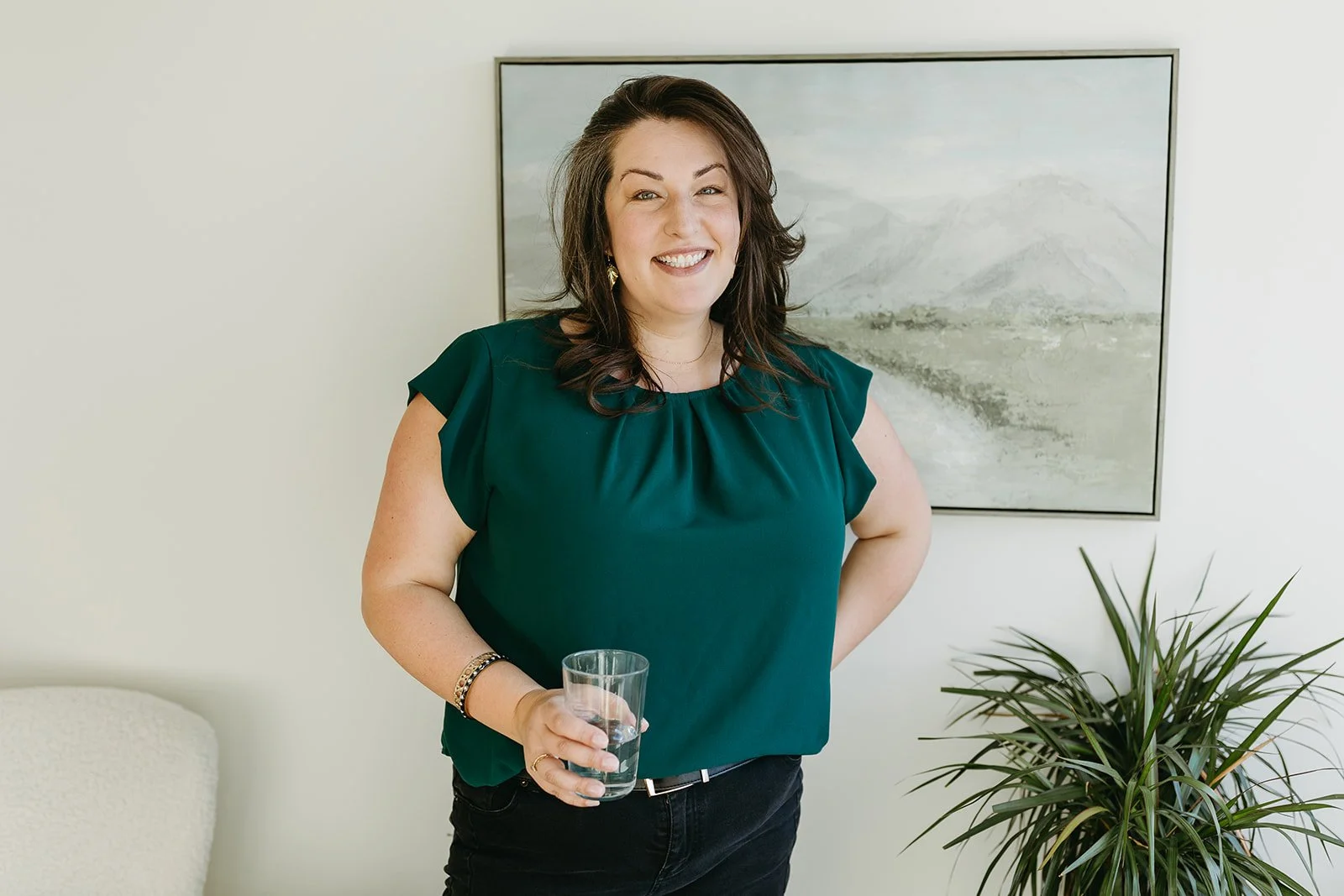 A woman with shoulder-length brown hair, smiling and holding a glass of water, standing in a room with a light-colored wall, a framed landscape painting, and a potted plant.