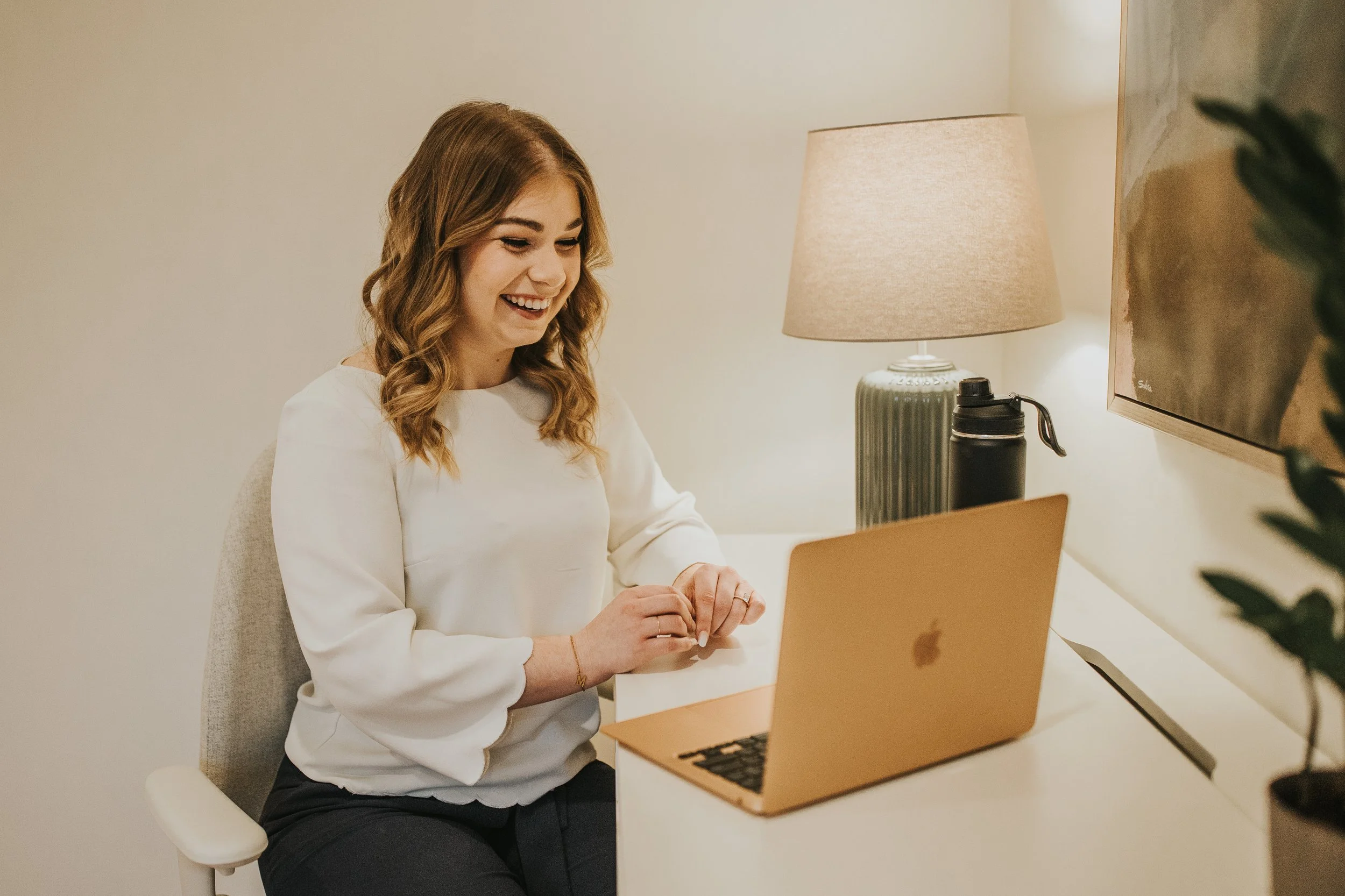 A woman with wavy brown hair smiling and sitting at a desk using a gold MacBook laptop, with a lamp and a water bottle on the desk near a wall-mounted TV.