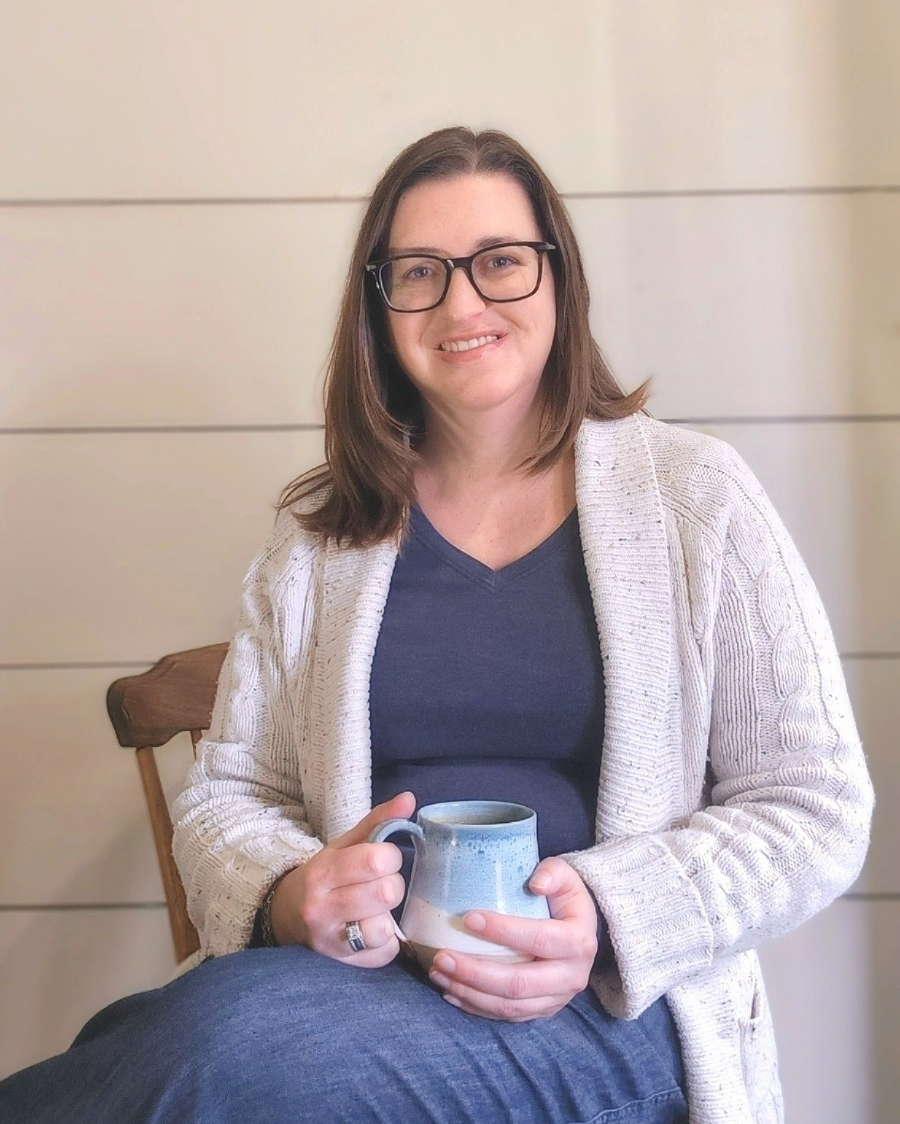 A woman with glasses and brown hair smiling, sitting on a wooden chair, holding a coffee mug, wearing a navy shirt and a light cardigan, against a neutral beige wall.