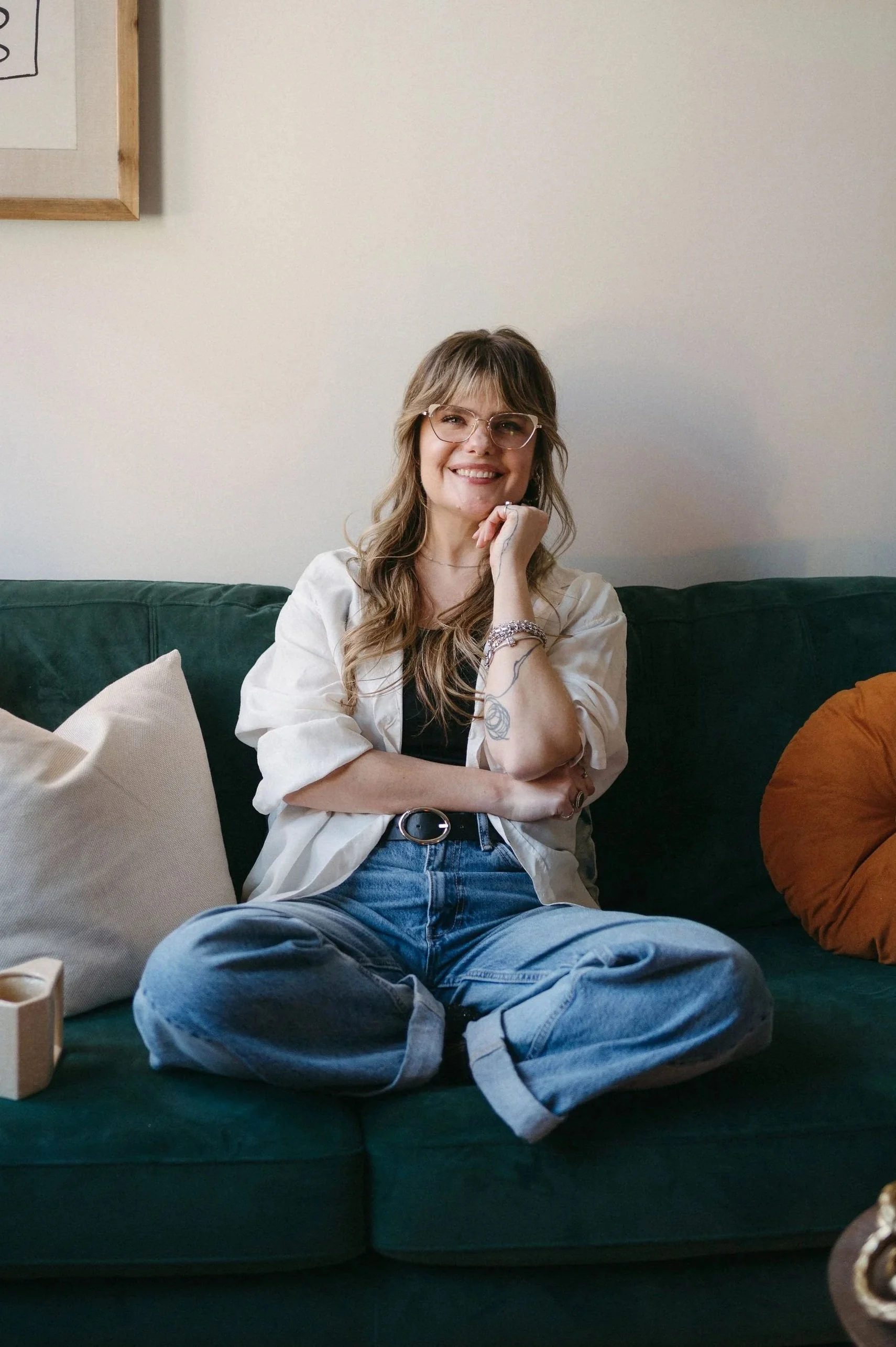 A woman sitting cross-legged on a green couch smiling at the camera, wearing glasses, a white shirt, black top, and jeans with rolled-up cuffs, with pillows around her and a framed picture on the wall behind.