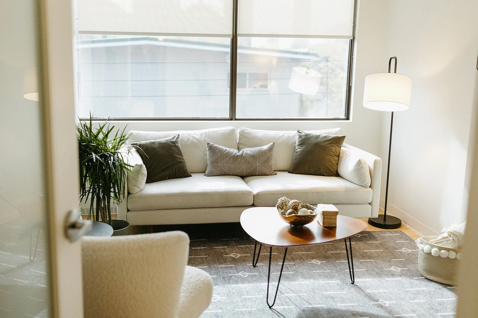 Living room with a white sofa, dark throw pillows, a round wooden coffee table, a potted plant, a floor lamp, and a large window.