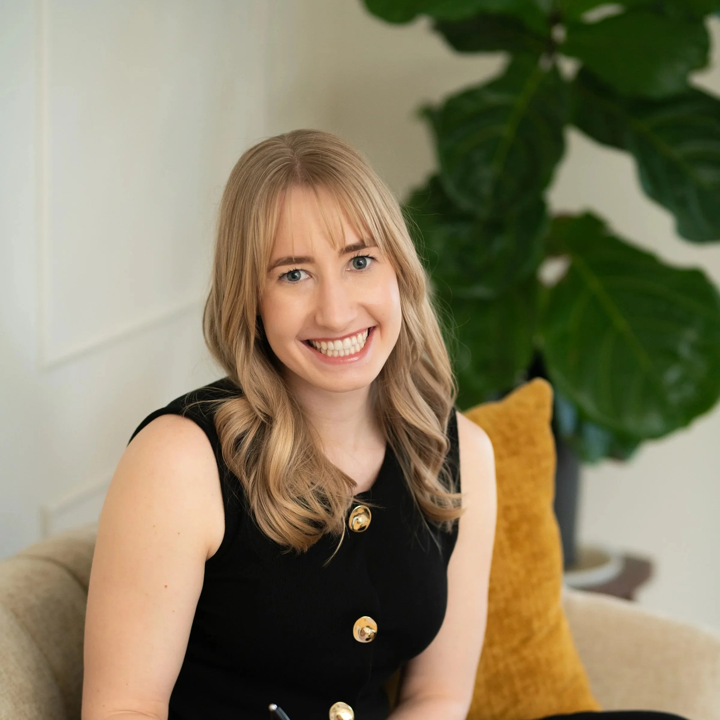 A woman with long blonde hair wearing a black vest with gold buttons and black pants sitting on a beige sofa with a green plant in the background. She is smiling at the camera and holding a notebook and pen.