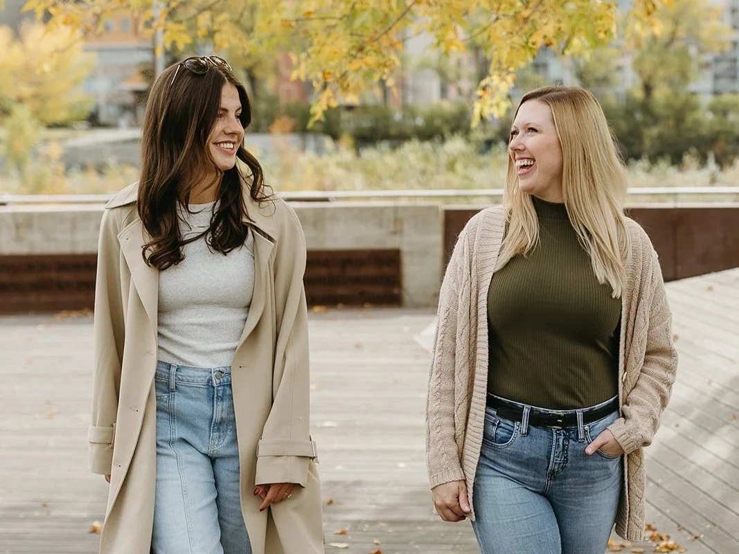 Two women are smiling and talking outdoors on a fall day, with trees and a park in the background.
