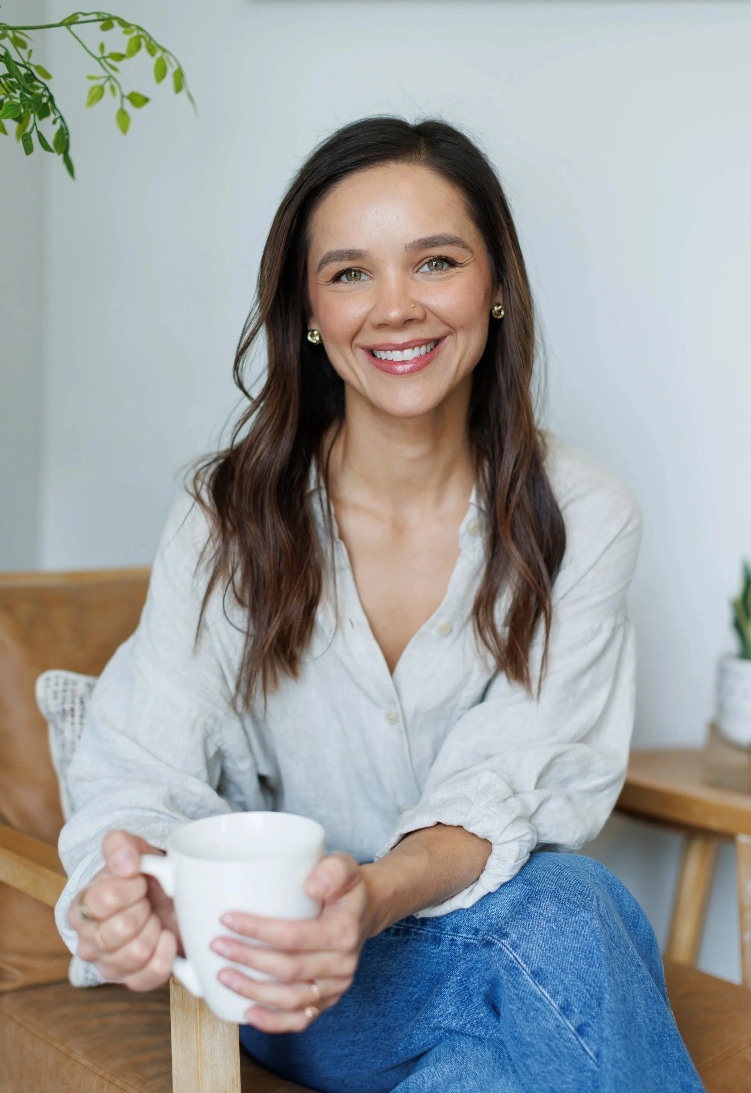 A young woman with long brown hair, sitting in a wooden chair holding a white mug